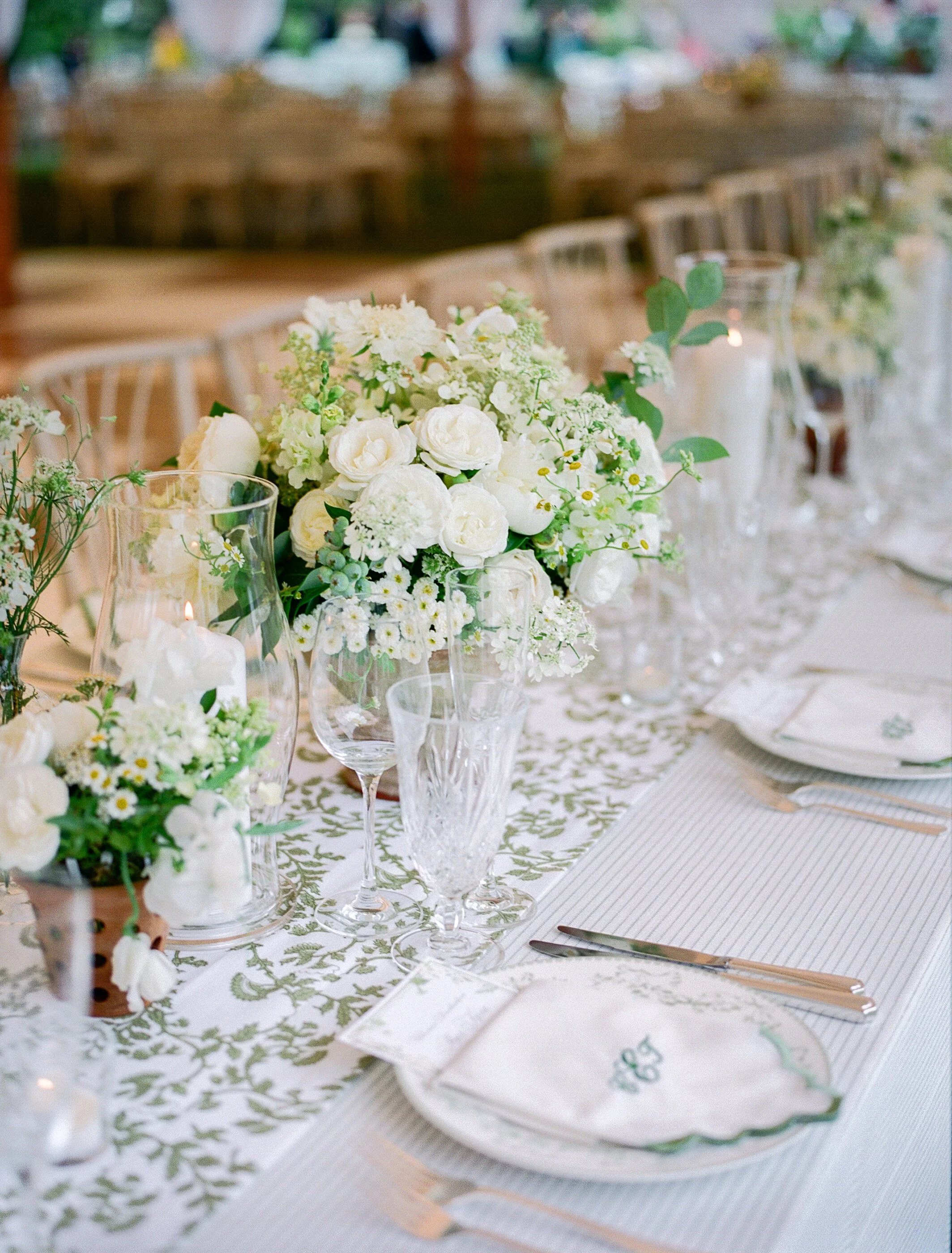 Elegant wedding table with white floral centerpiece, candles, glassware, and fine china on a decorated tablecloth.