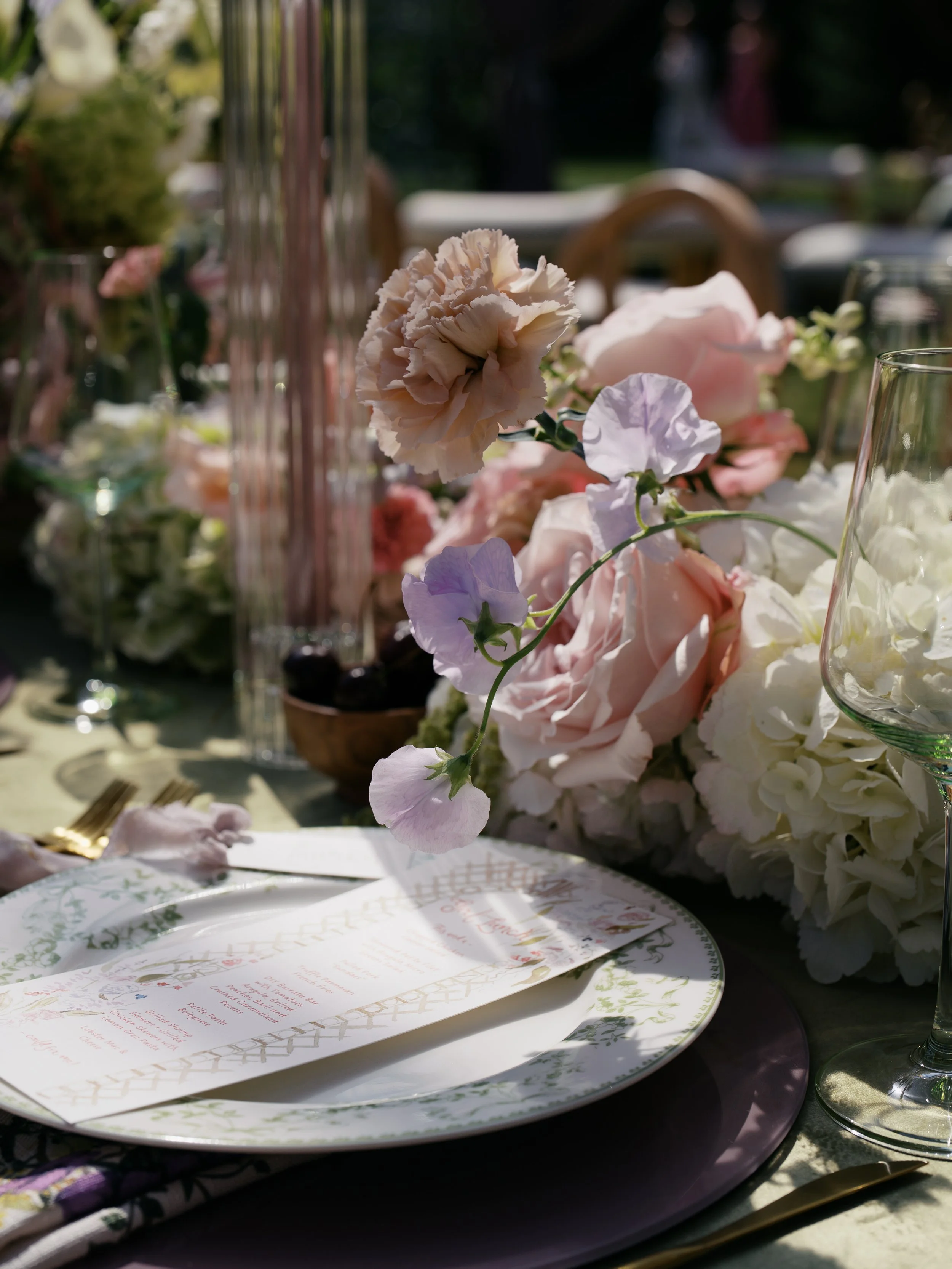 Close-up of a beautifully set outdoor dining table with floral arrangements, elegant tableware, and sunlight.