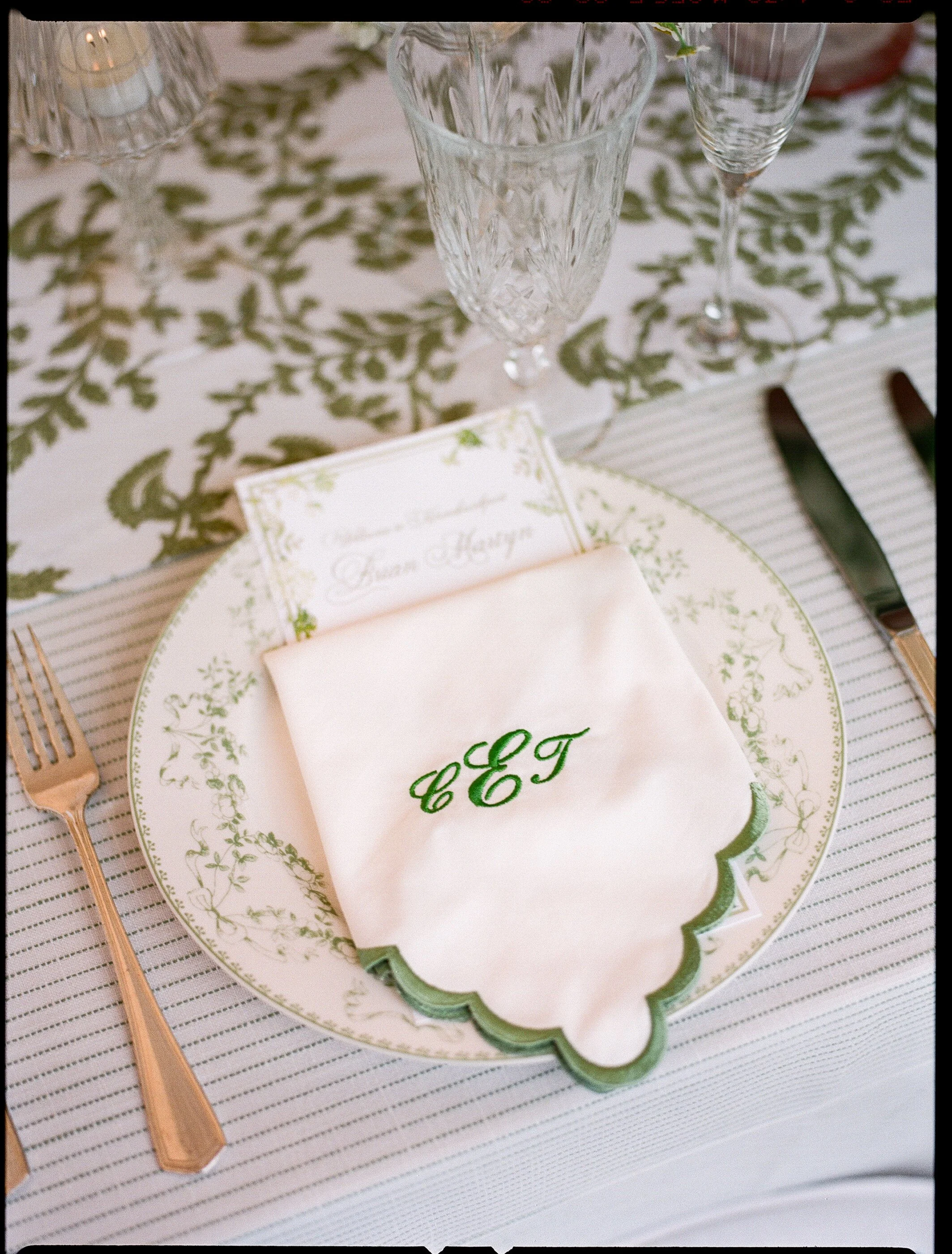 A formal table setting with a decorative plate, a napkin with green initials, a gold fork, and crystal glasses on a tablecloth with a floral and striped pattern.