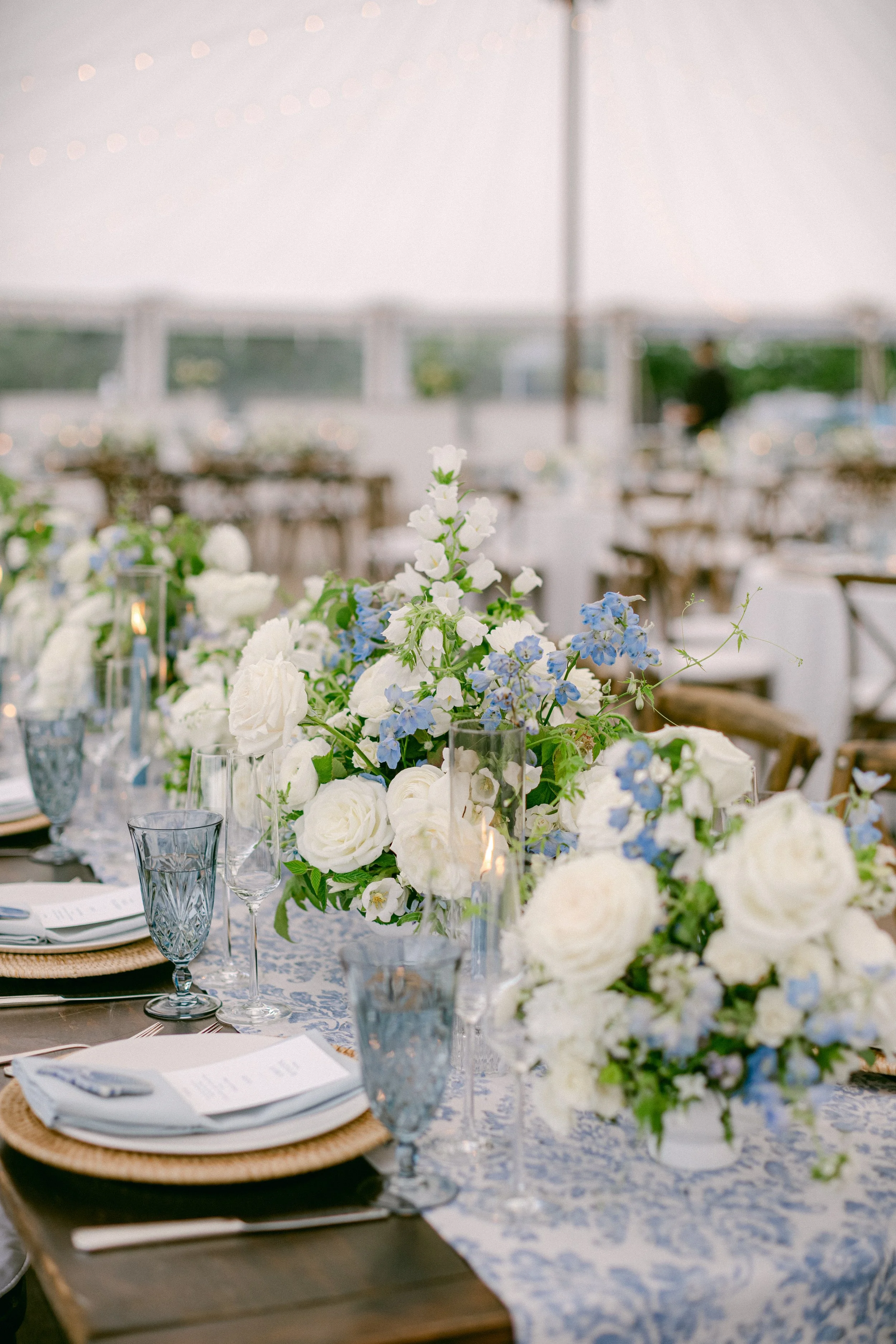 Elegant outdoor table decorated with white and blue floral arrangements, candles, and place settings for a wedding or special event, under a white tent.
