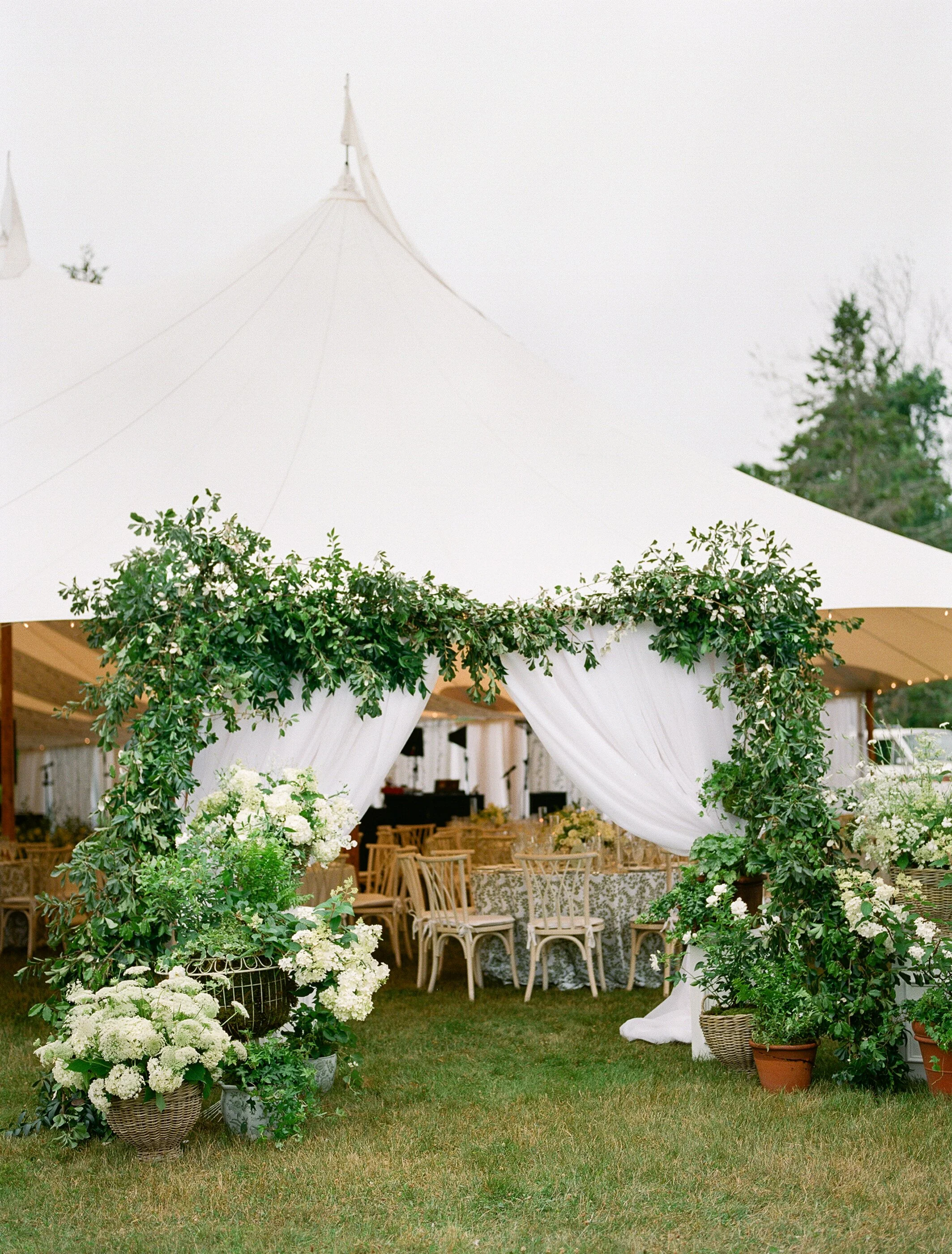 Outdoor wedding tent decorated with greenery and white flowers, with wooden chairs and tables inside.