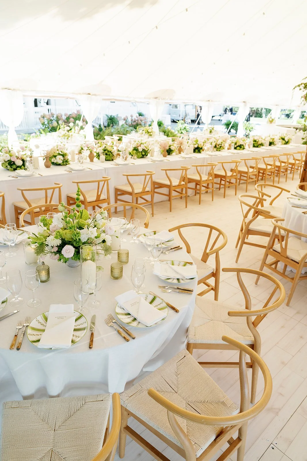 Elegant reception tent with long head table and round tables decorated with white linens, floral centerpieces, and candles, set for a wedding or event.