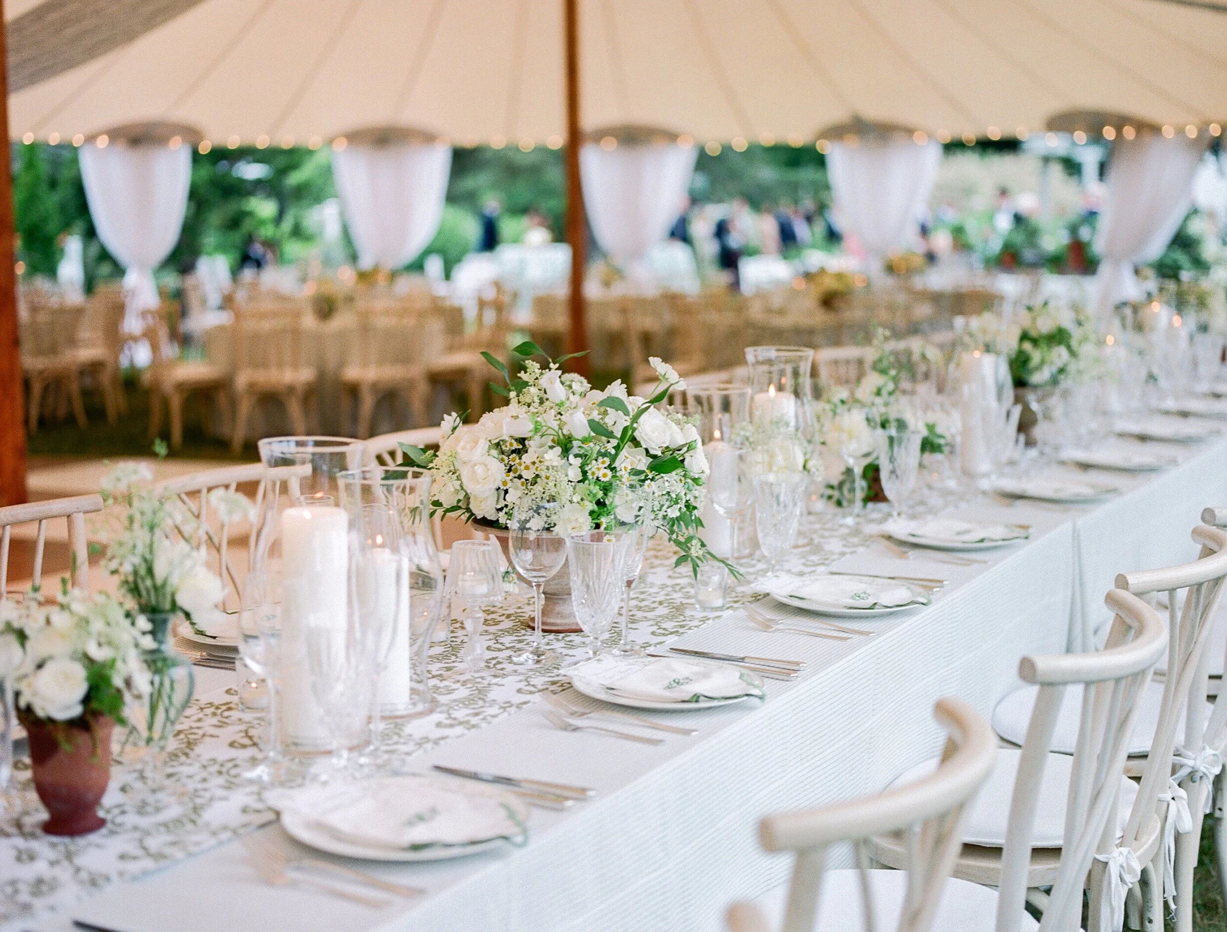 Elegant outdoor wedding reception under a tent with long table setup, white floral centerpieces, candles, and white chairs.