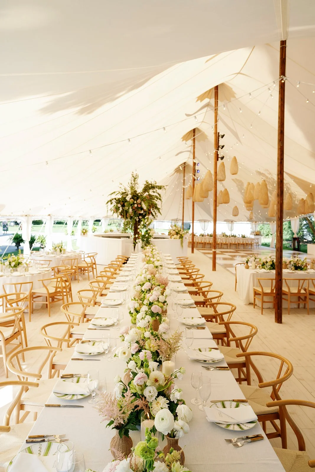 Elegant wedding reception tent with long table decorated with pink and white flowers, surrounded by wooden chairs, with smaller tables and large floral arrangements in the background.