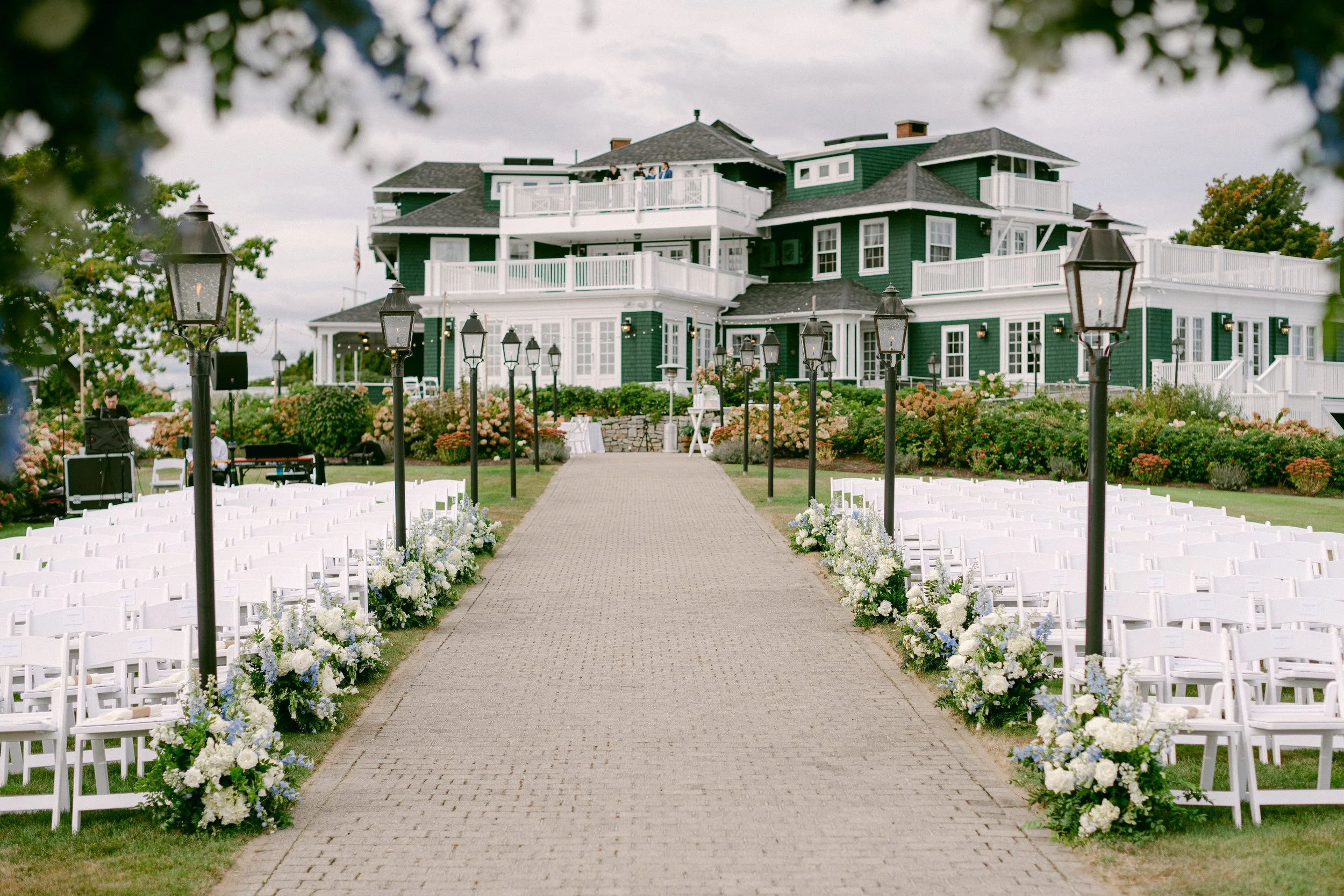 Outdoor wedding ceremony setup with white chairs and floral arrangements along a paved aisle, hotel or large house in the background, with black lamp posts lining the aisle, overcast sky.