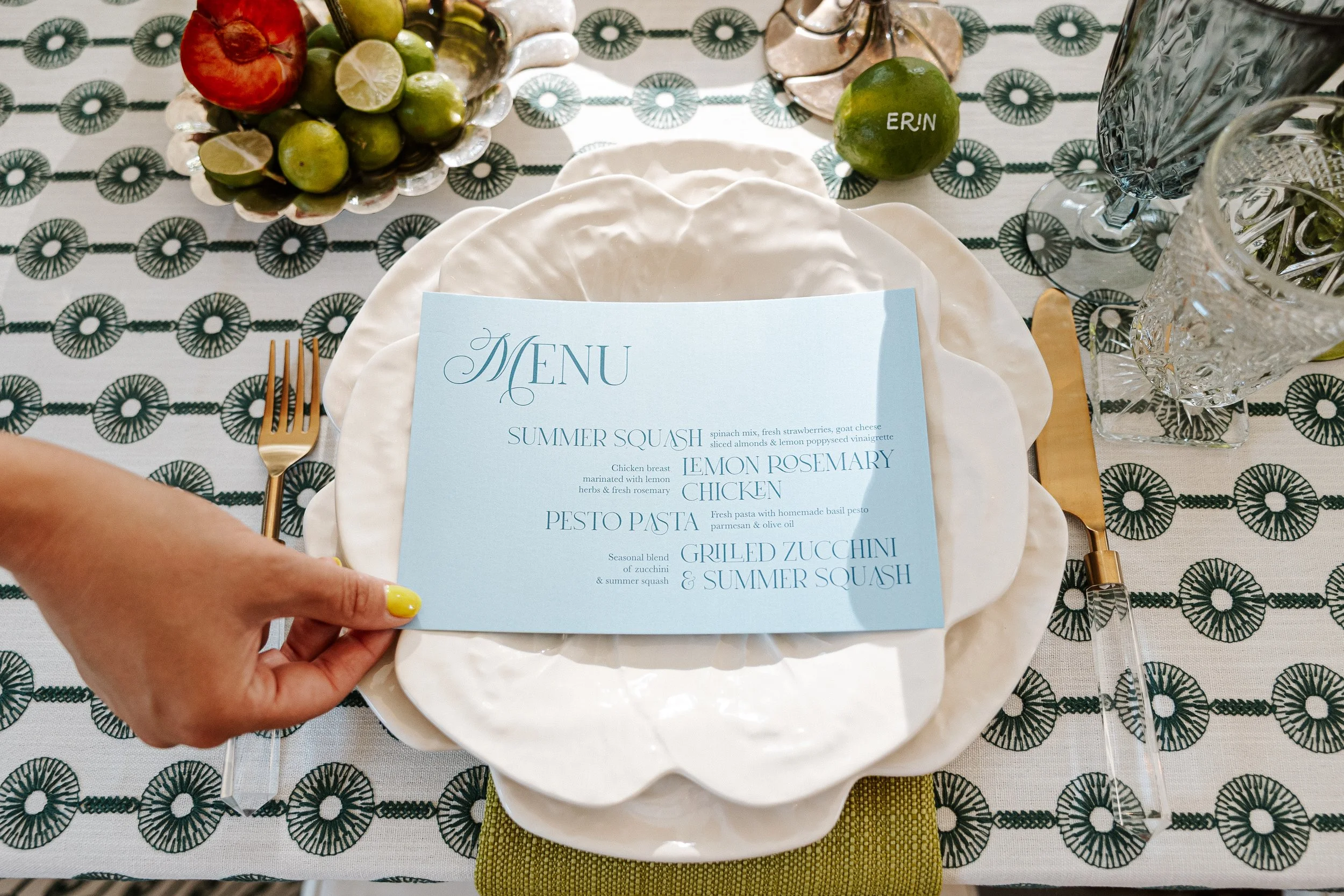 Table setting with a light blue menu card on a white plate, gold cutlery, a bowl of limes and an apple, and decorative glassware, with a patterned tablecloth.
