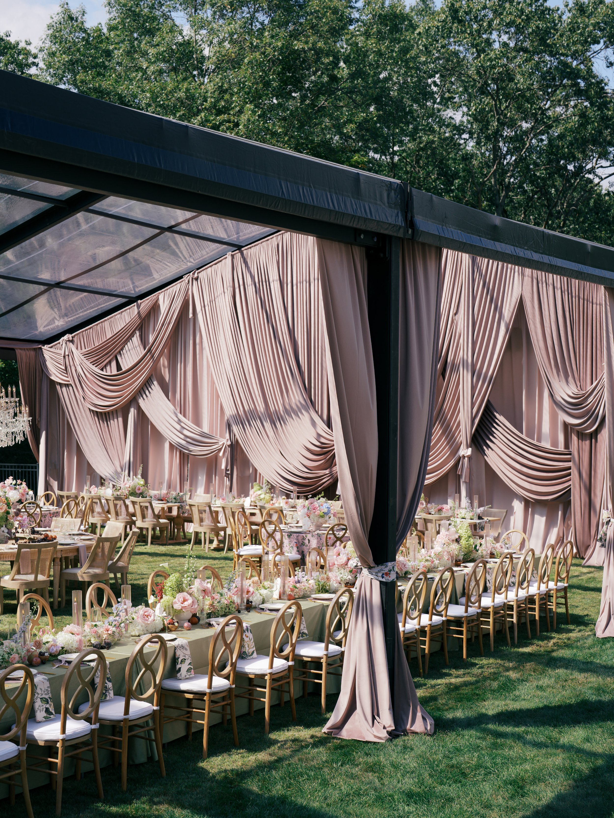 Elegant outdoor wedding setup with long tables adorned with pink and white floral arrangements, under a large pink draped tent with black support structure, surrounded by greenery.