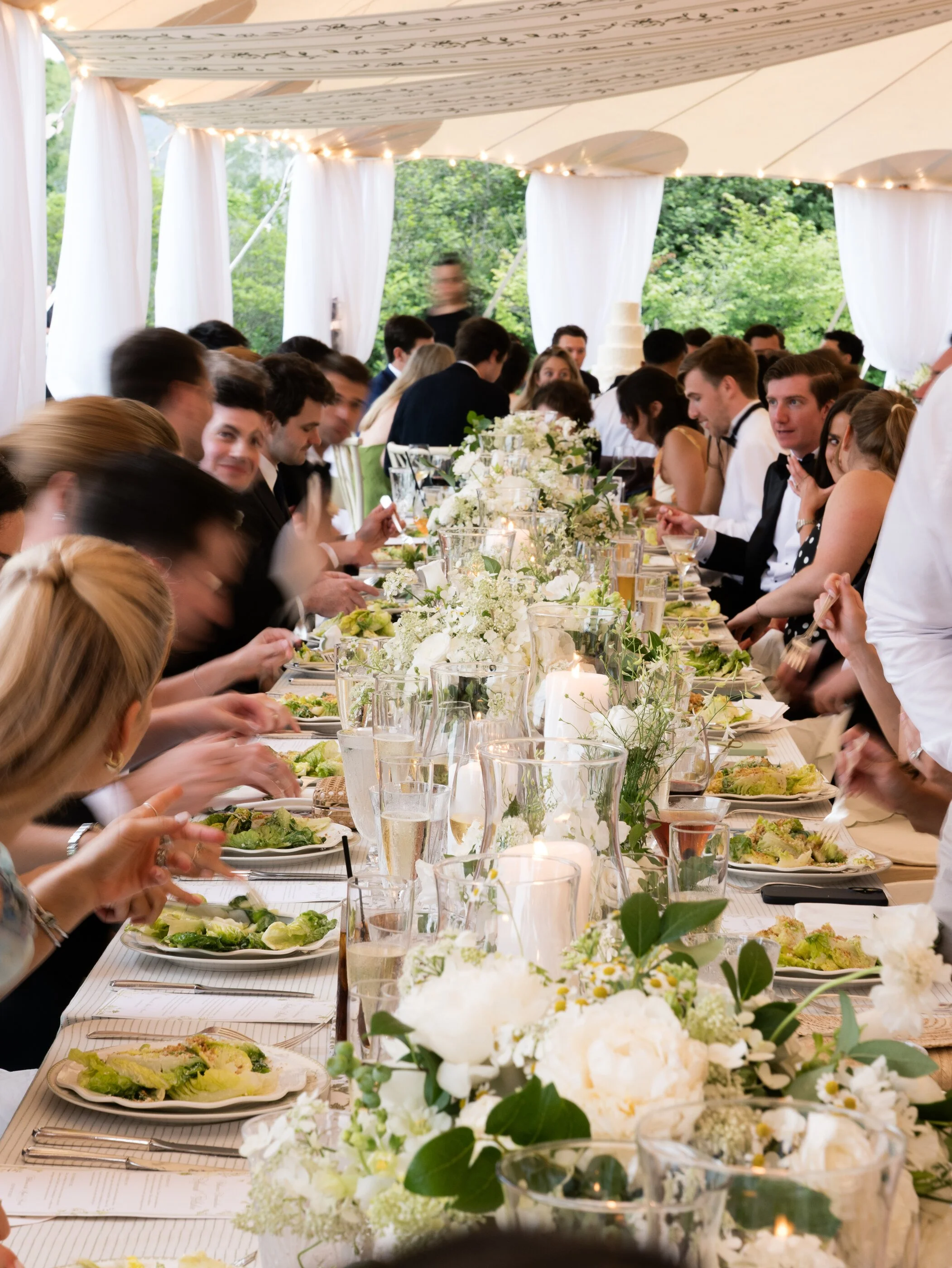 A large outdoor wedding reception under a white tent with long tables decorated with white flowers and candles. Guests are seated on both sides, enjoying their meal and engaging in conversation.