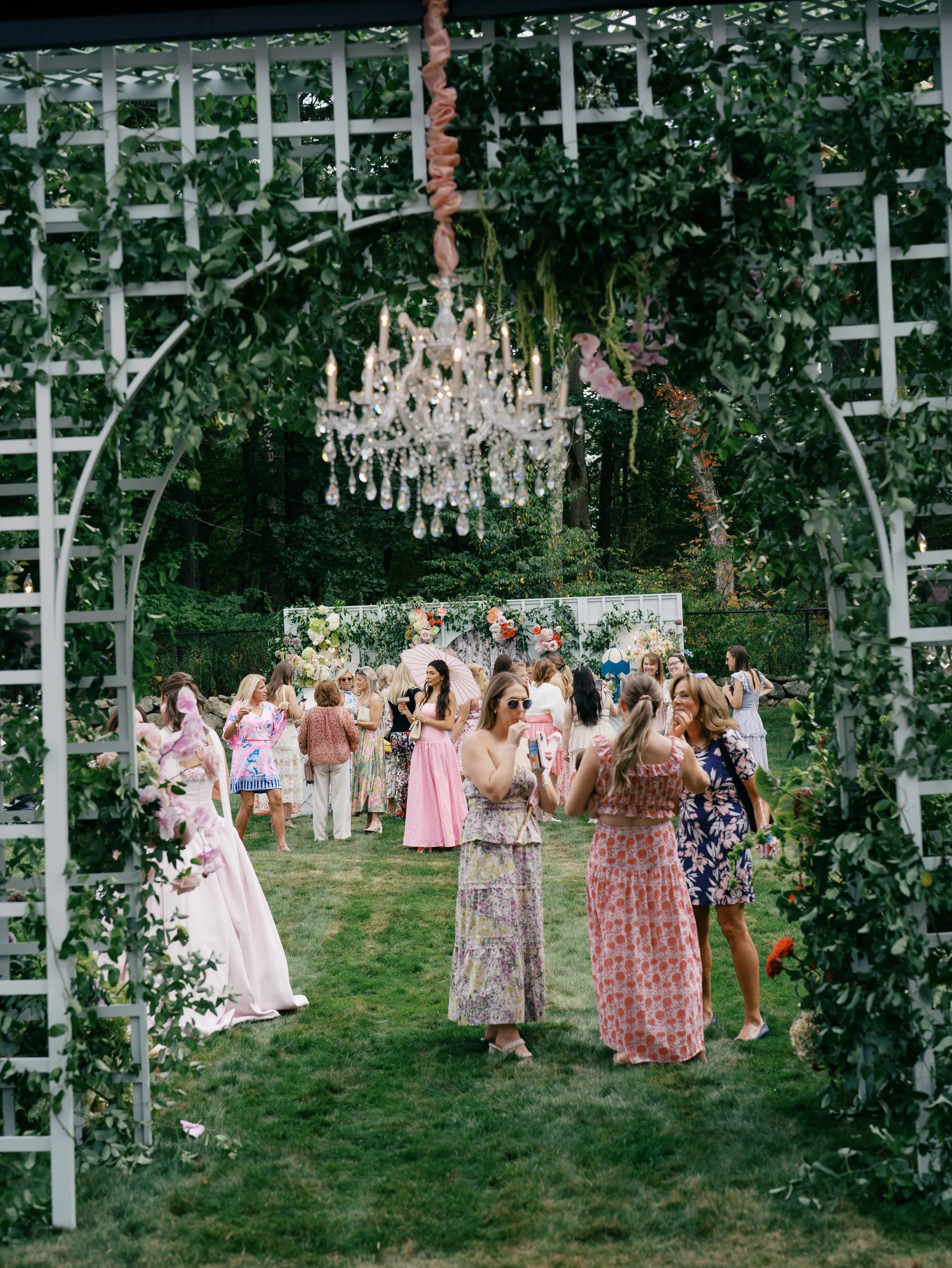 People at a garden party seen through a decorative white garden arch with a hanging chandelier at the top. The guests are mostly women in colorful summer dresses, standing and chatting on a lawn with a floral backdrop.