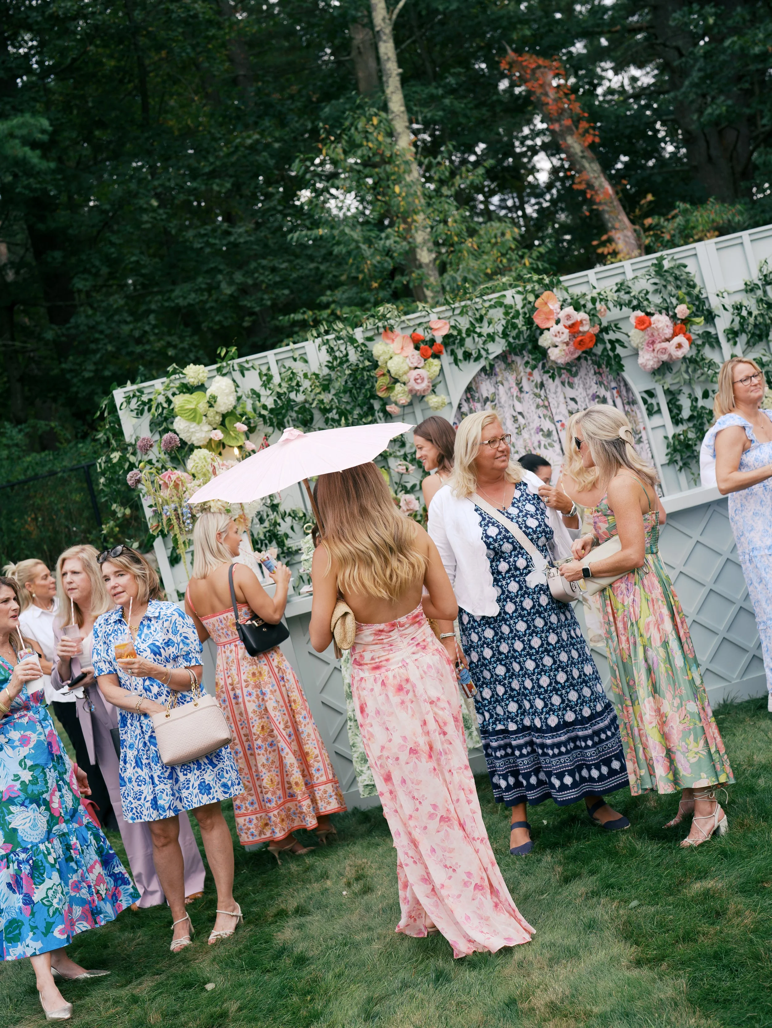 Group of women in floral dresses socializing at outdoor garden party with floral backdrop