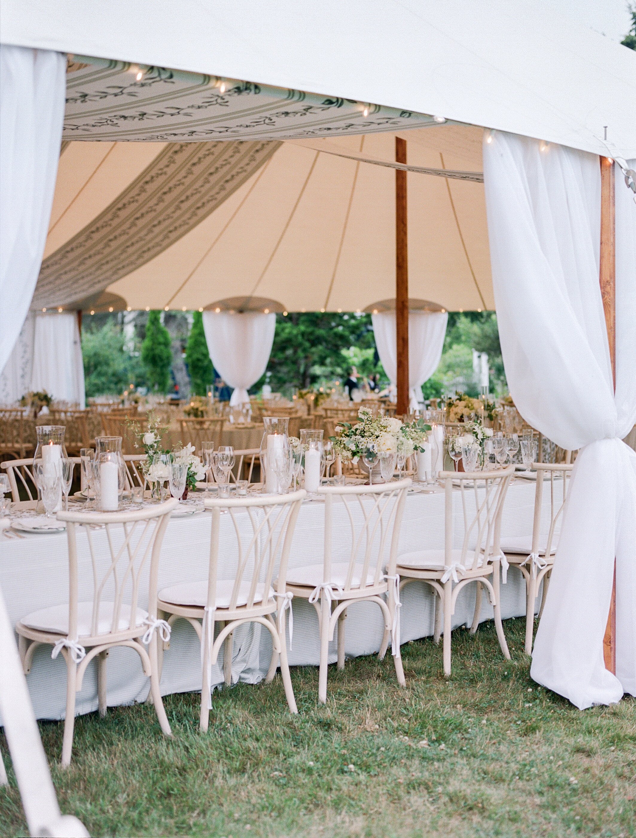 Elegant outdoor wedding reception under a white canopy tent with tables decorated with white and green floral centerpieces, candles, and glassware. Wooden chairs with tied bows surround the tables, and the scene is set on a grassy area with trees in 