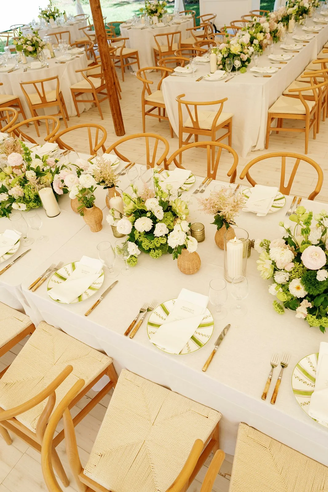 Elegant wedding reception table decorated with white tablecloths, floral centerpieces in white, blush, and green, candles, and place settings with plates, silverware, napkins, and glasses.