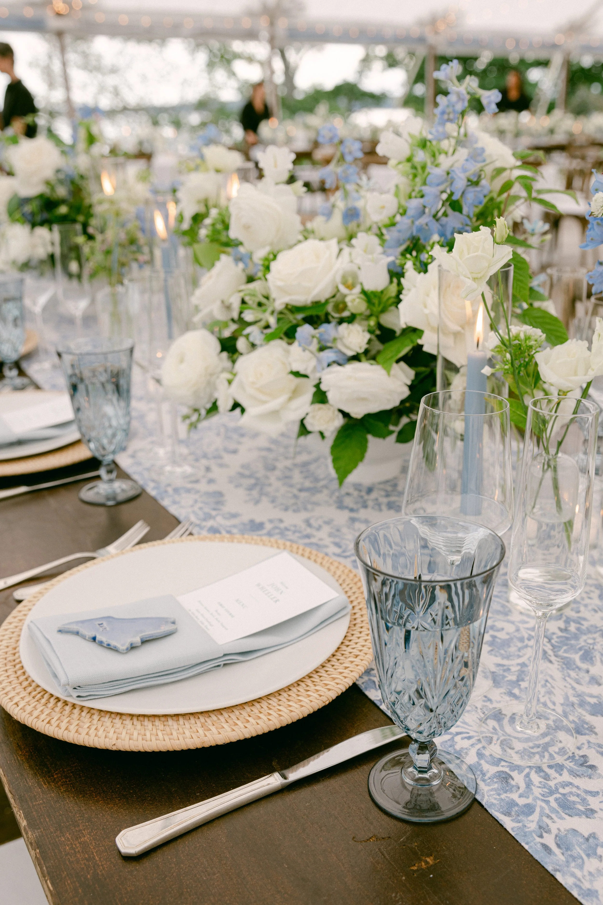 Table setting at an outdoor event with a floral centerpiece of white and blue flowers, glassware, plates, napkins, and a blue patterned table runner.