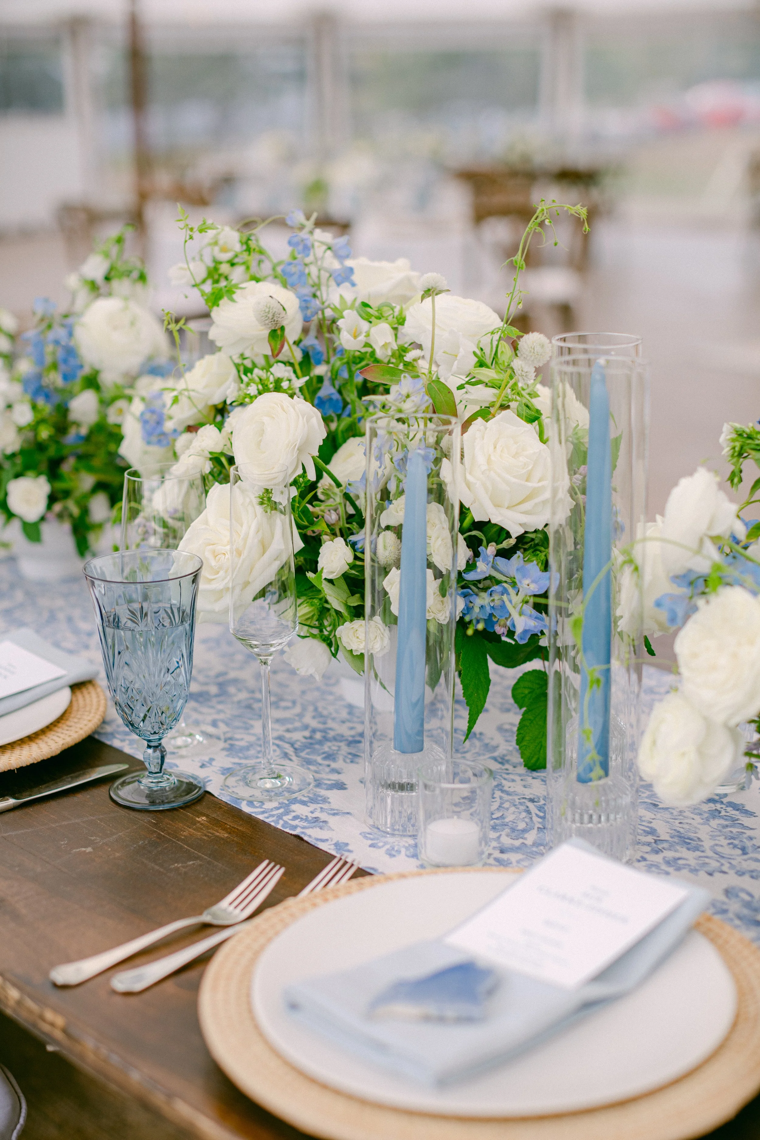 Table setting with white and blue floral centerpiece, blue candles, place setting with a menu, and elegant glassware at an event or wedding.
