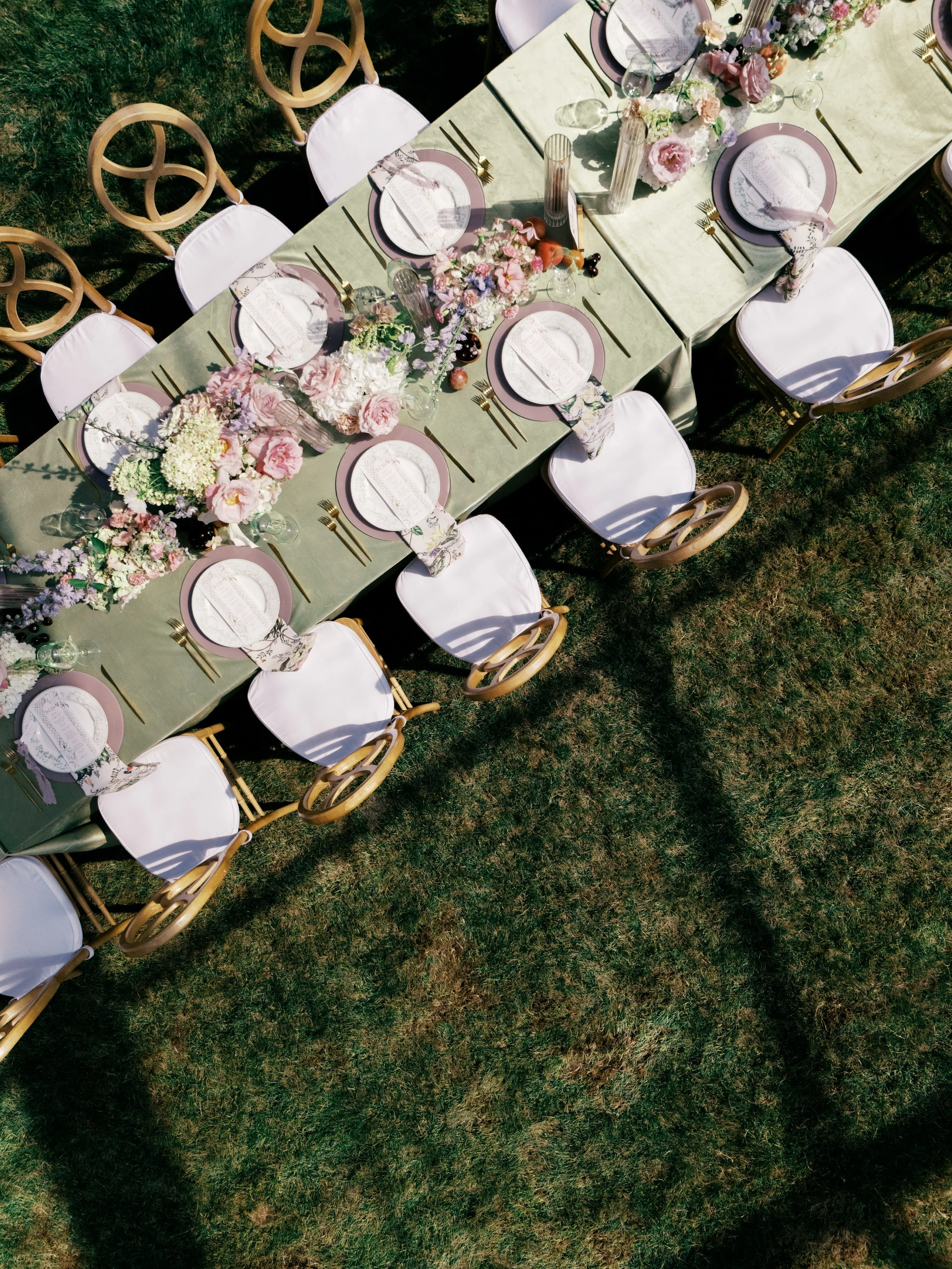 Top-down view of an outdoor banquet table set with pink and white floral arrangements, plates, gold flatware, and white chairs on grass.