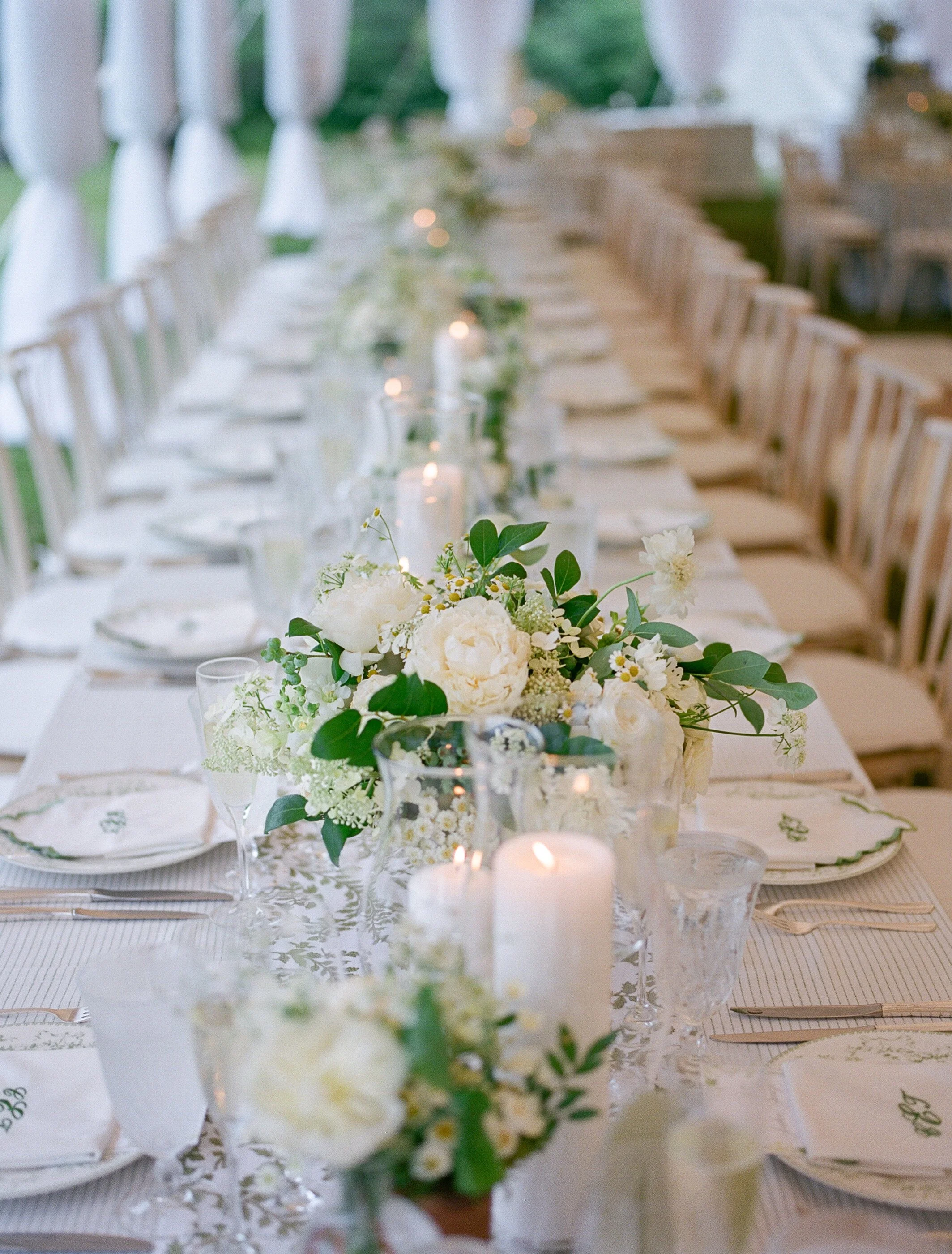 Long dining table set for an event with white floral arrangements, candles, and elegant place settings in an outdoor tent.