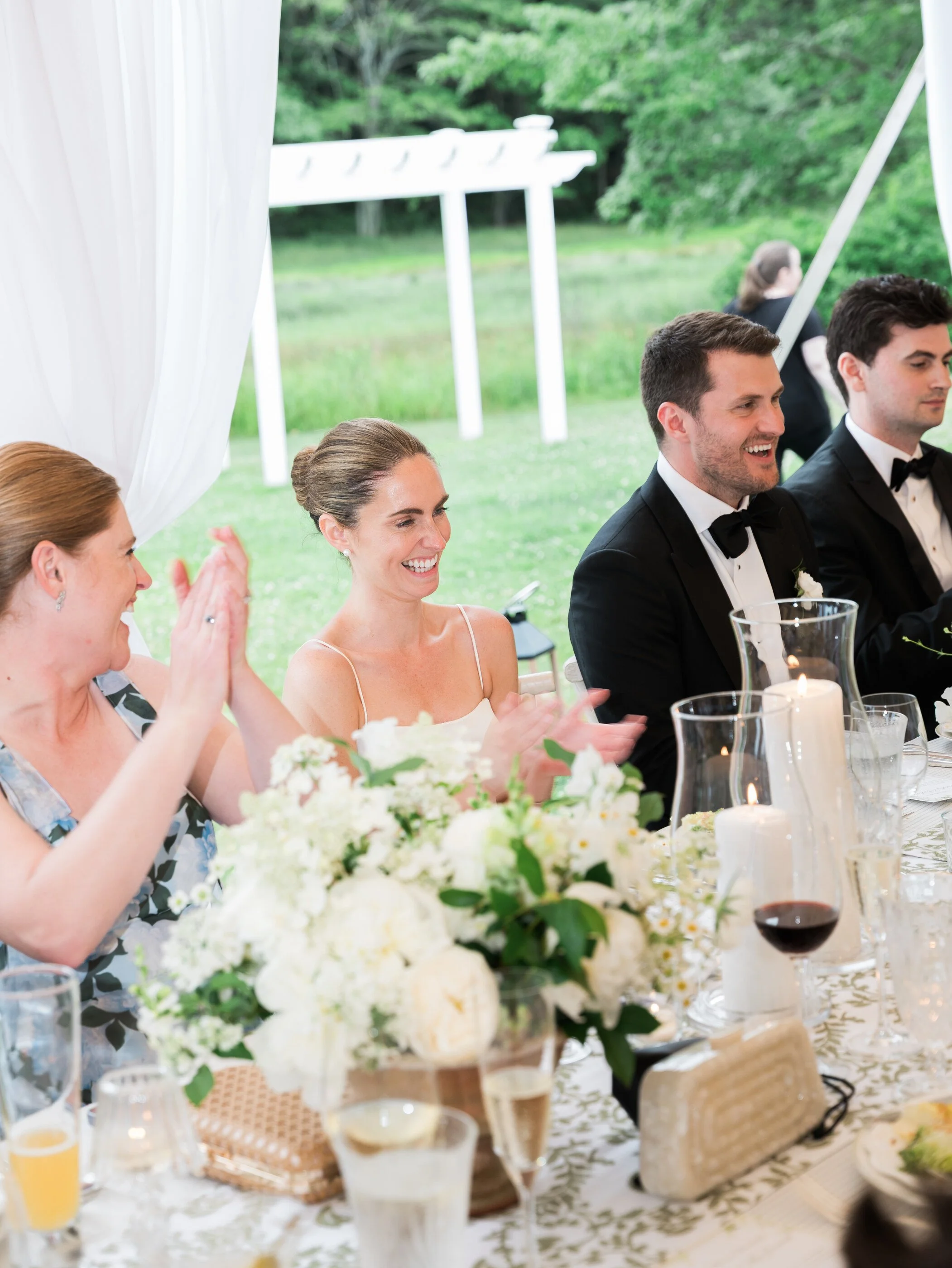 People at a wedding reception sitting at a decorated table, clapping and smiling, outdoors with greenery and a white tent or pavilion in the background.