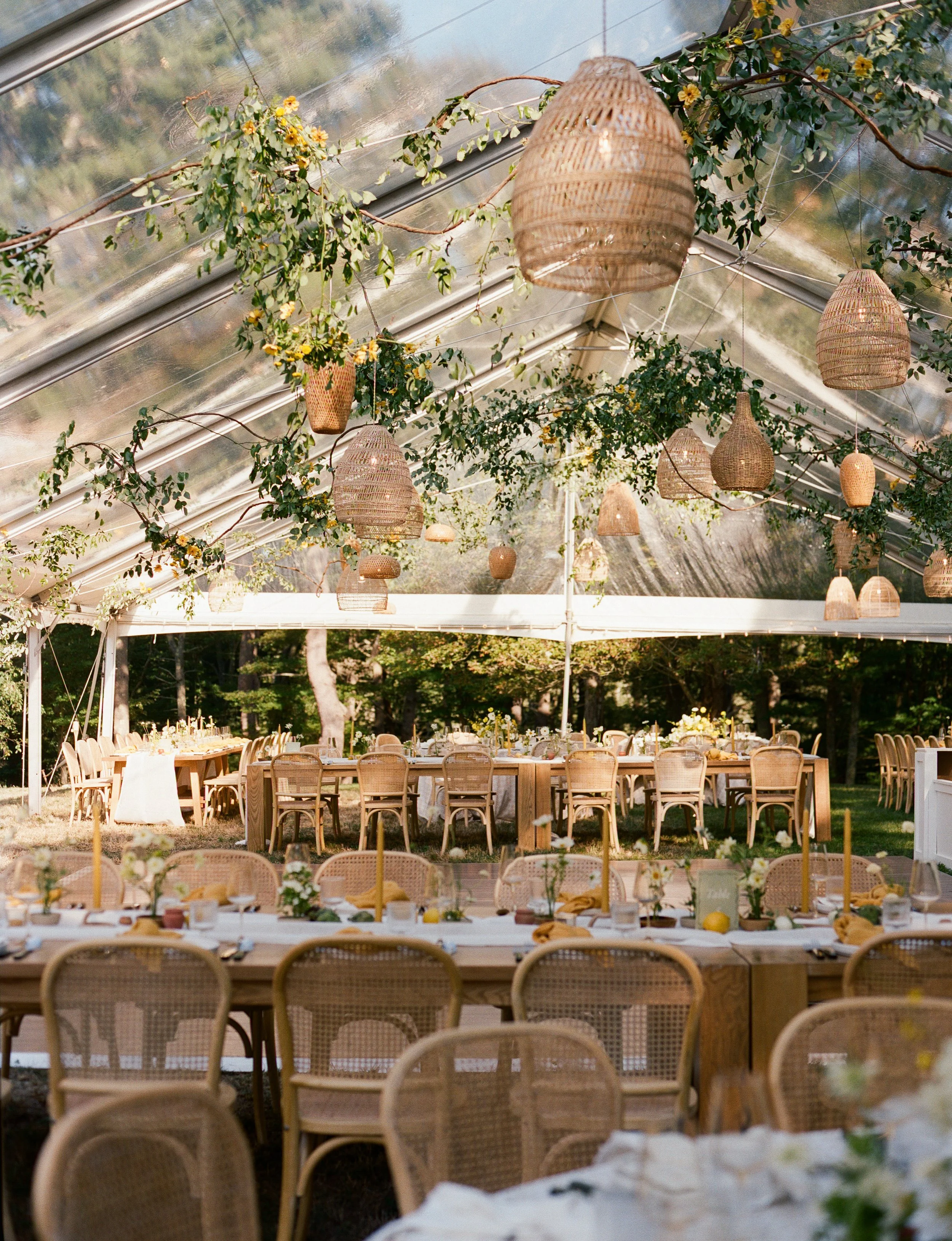 Decorated outdoor wedding or event reception under a transparent tent, with hanging wicker lanterns and lush greenery, wooden tables, and rattan chairs.