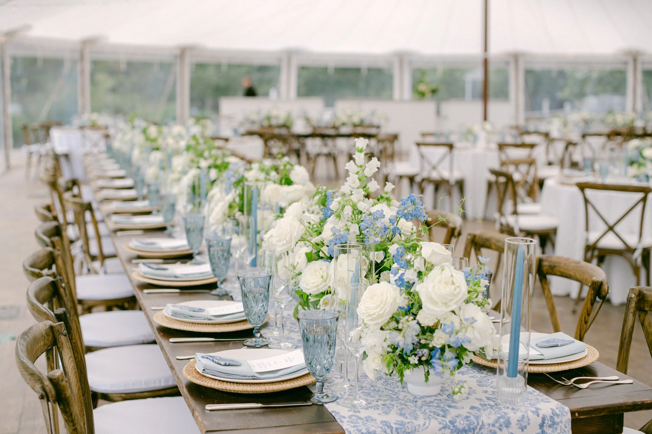Elegant outdoor wedding or event table setup with white and blue floral centerpieces, blue glassware, white plates, and wooden chairs under a tent.