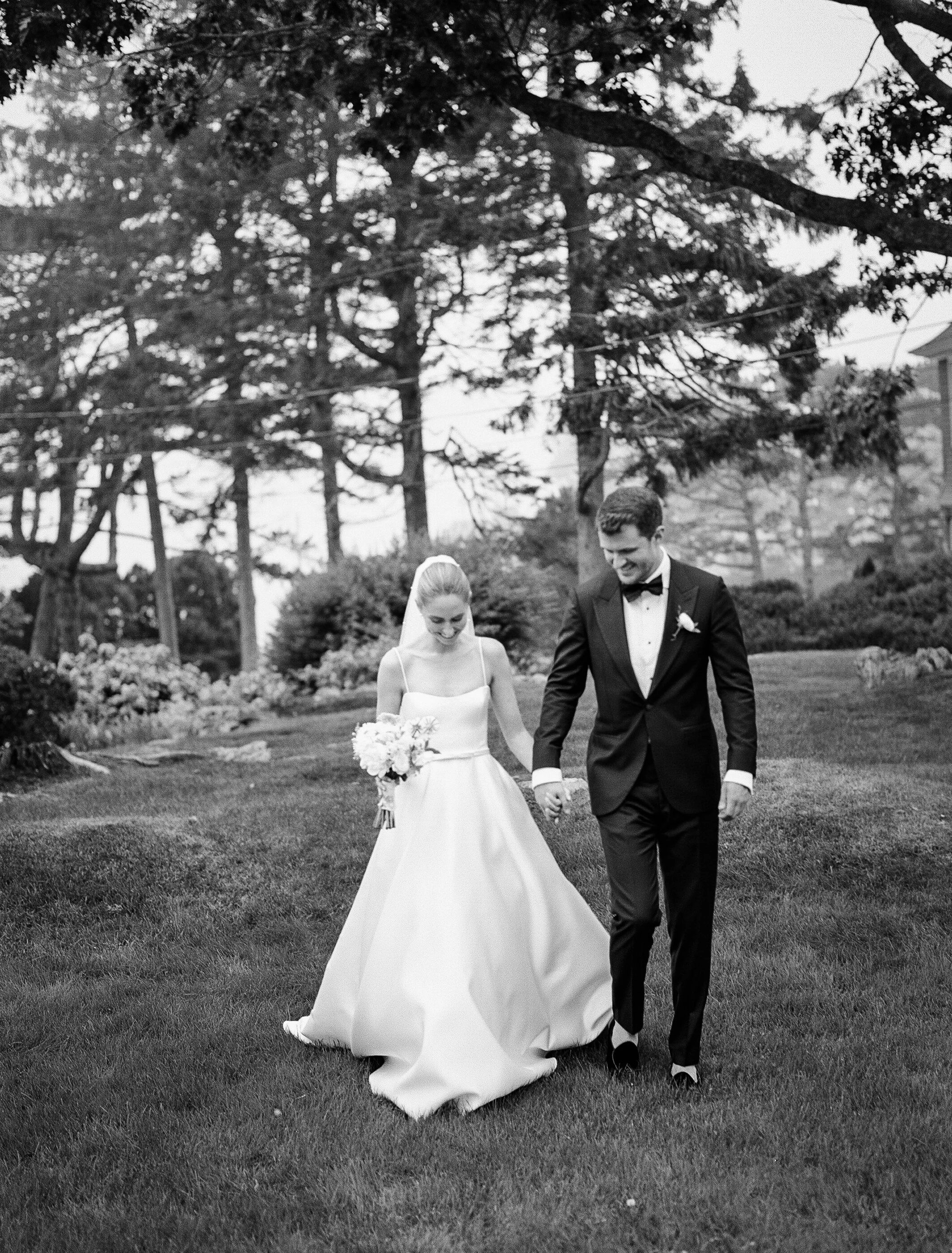 A bride and groom walking hand in hand outdoors on a wedding day, smiling, with trees and bushes in the background, in black and white.