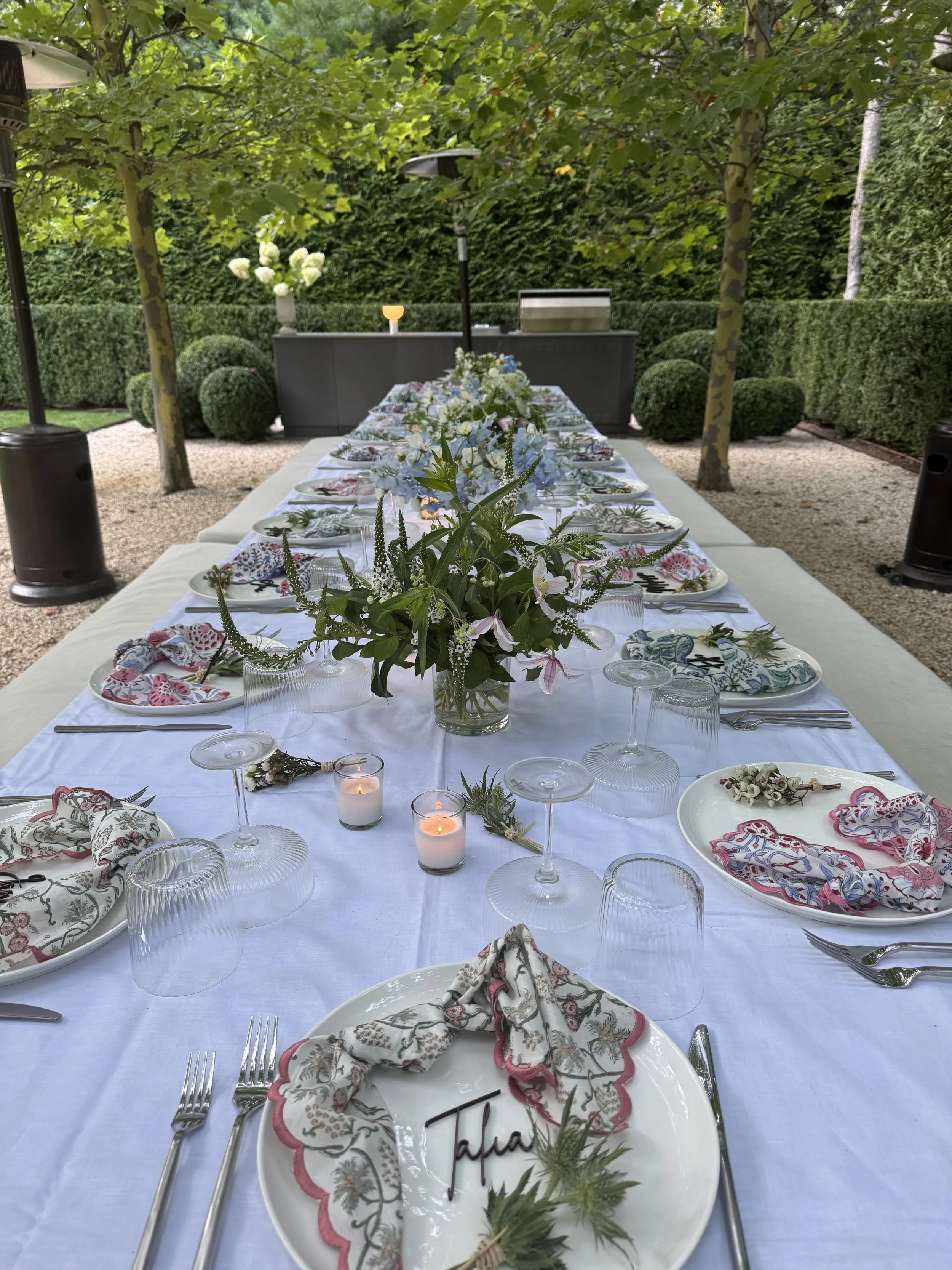 Outdoor formal dining table set with white tablecloth, floral placemats, napkins, wine glasses, silverware, and a flower centerpiece. Lanterns and candles decorate the setting in a lush garden with trees, bushes, and hedges.