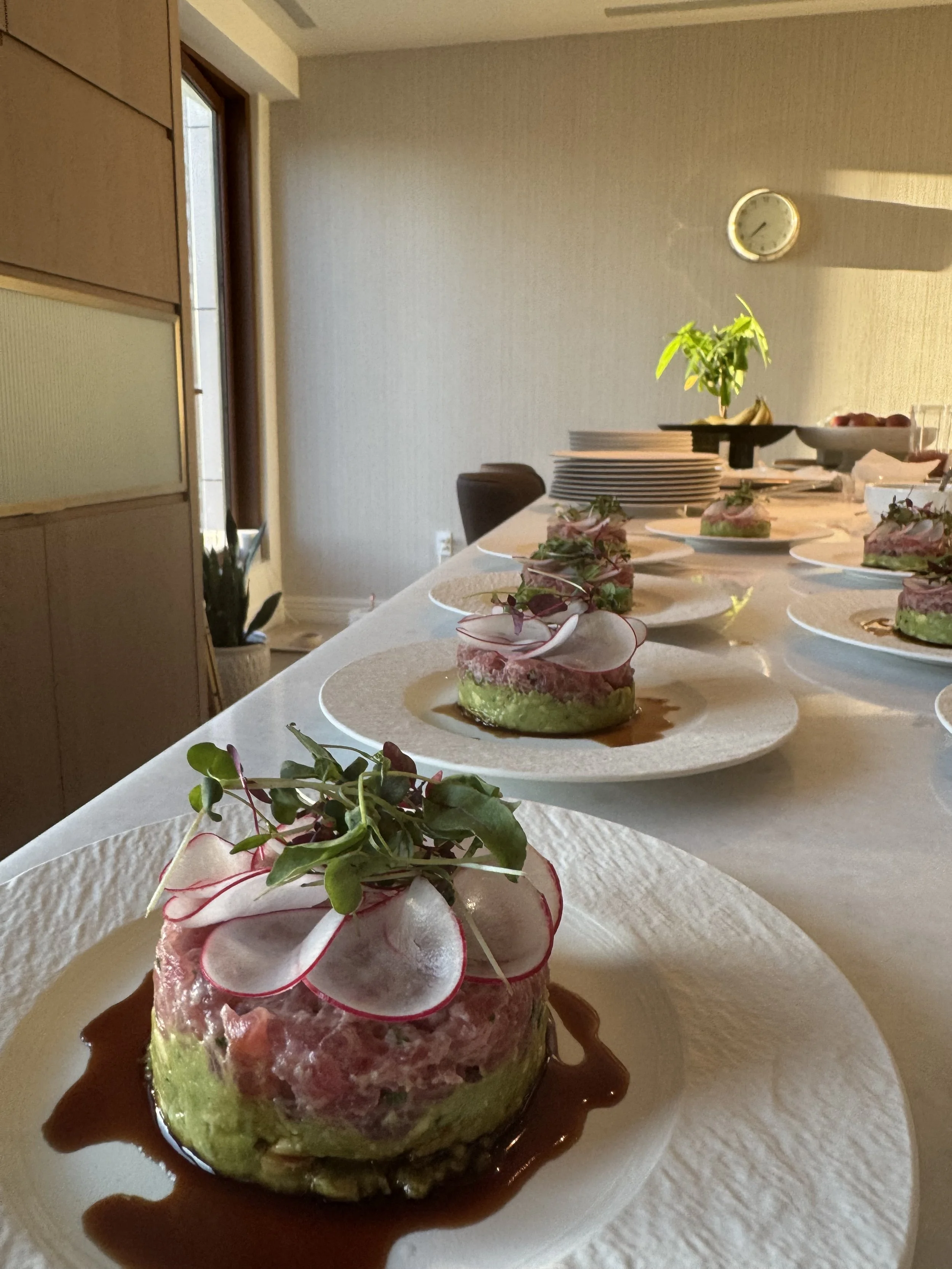 Close-up of a gourmet dish featuring a stack of layered ingredients garnished with microgreens on a white plate, with sauce drizzled around the base, inside a dining room with a table, chairs, a plant, and a wall clock.