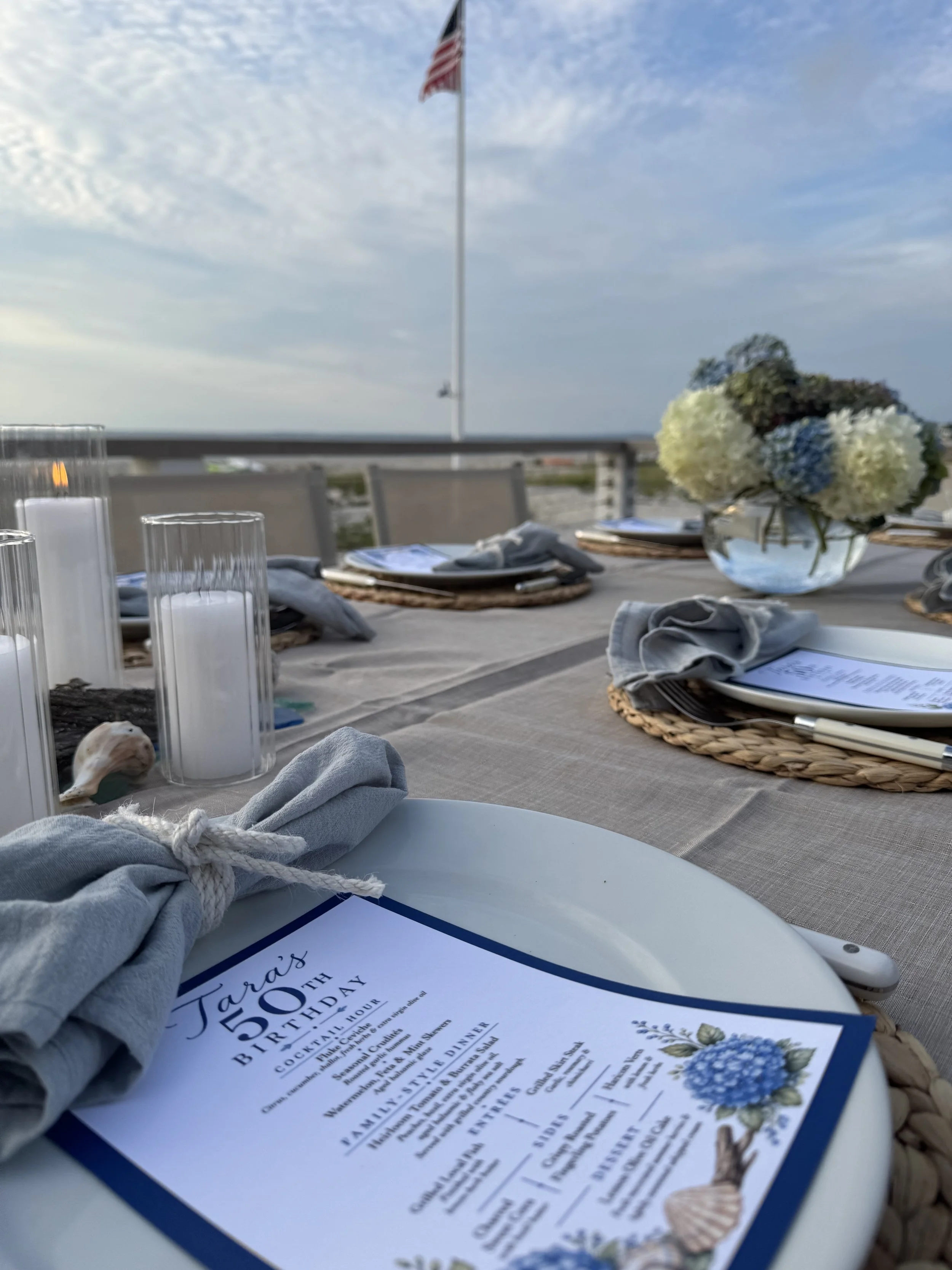 A table set for an outdoor celebration with a floral centerpiece, printed menus, candles, and cloth napkins, with an American flag hotel sign in the background and a partly cloudy sky.