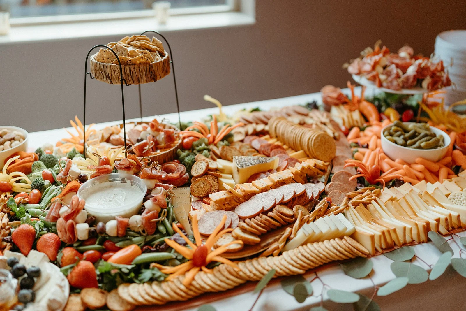 A large cheese and charcuterie board with various cheeses, meats, crackers, vegetables, fruits, and a bowl of green olives, decorated with orange flower-shaped vegetables and greenery.