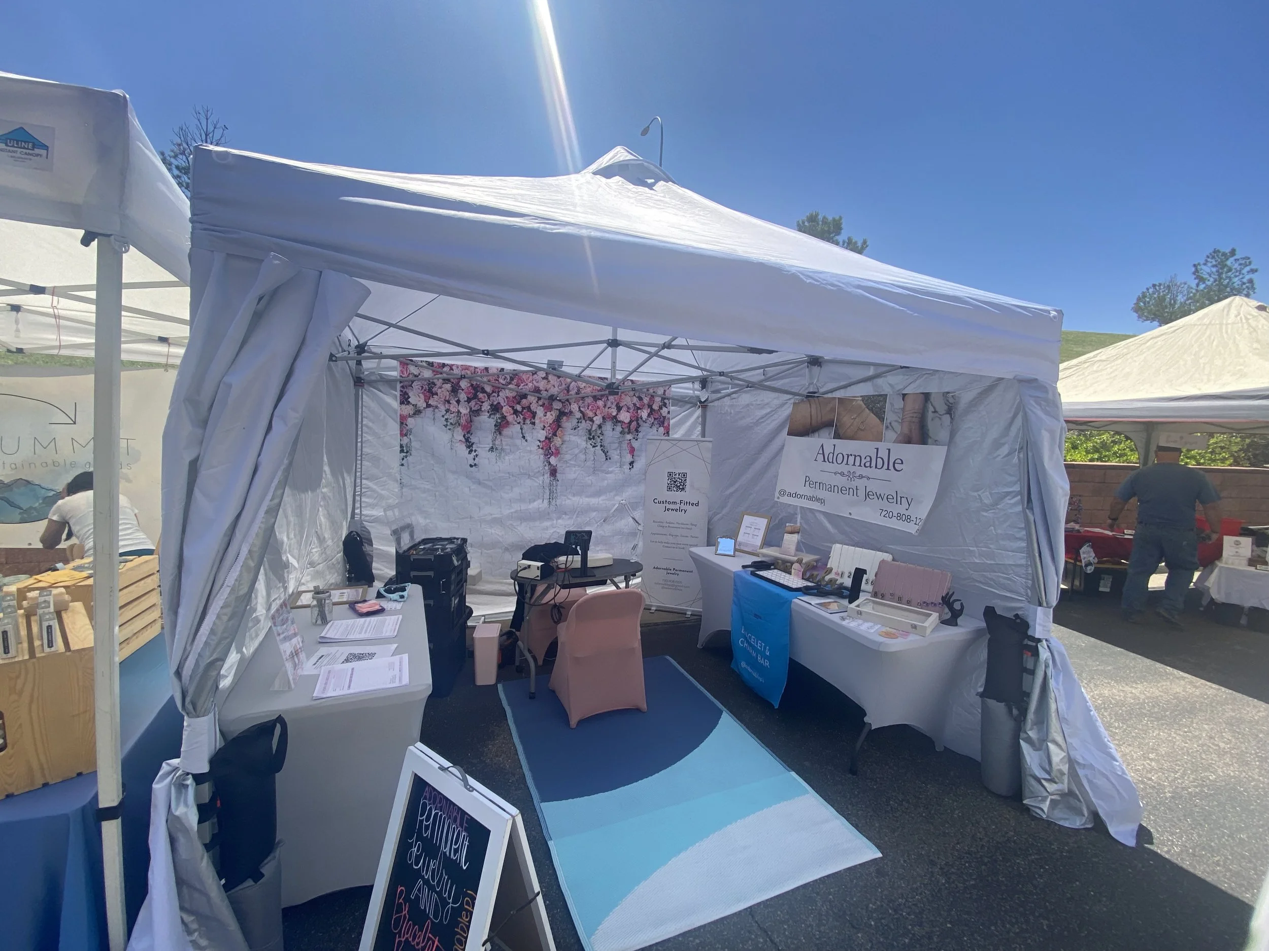 An outdoor jewelry booth with a white canopy tent, decorated with signs and jewelry displays, under a clear blue sky at a market or fair.
