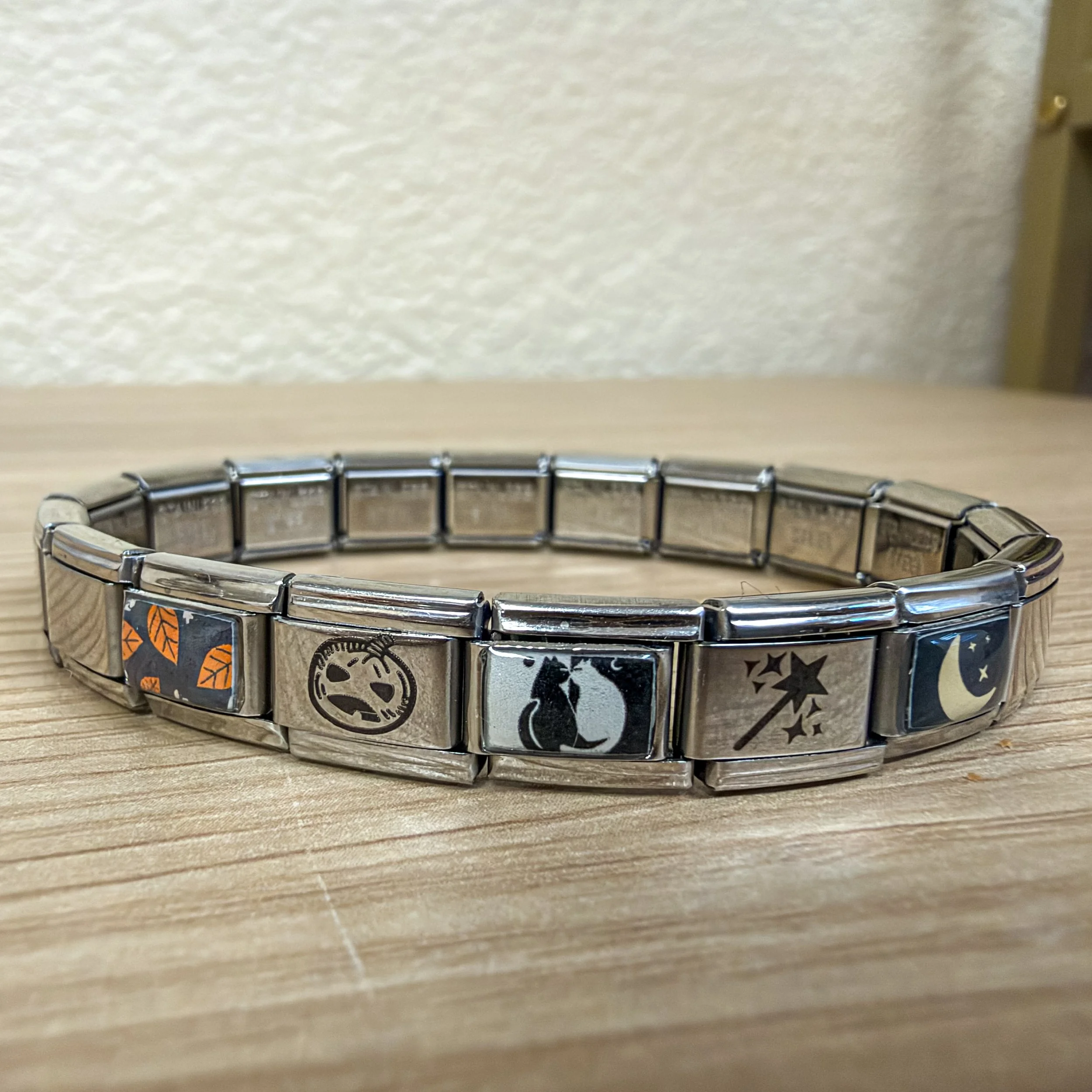 Silver Italian charm bracelet featuring five Halloween charms, including a pumpkin, black cats, magic wand, and a crescent moon, displayed on light wood.