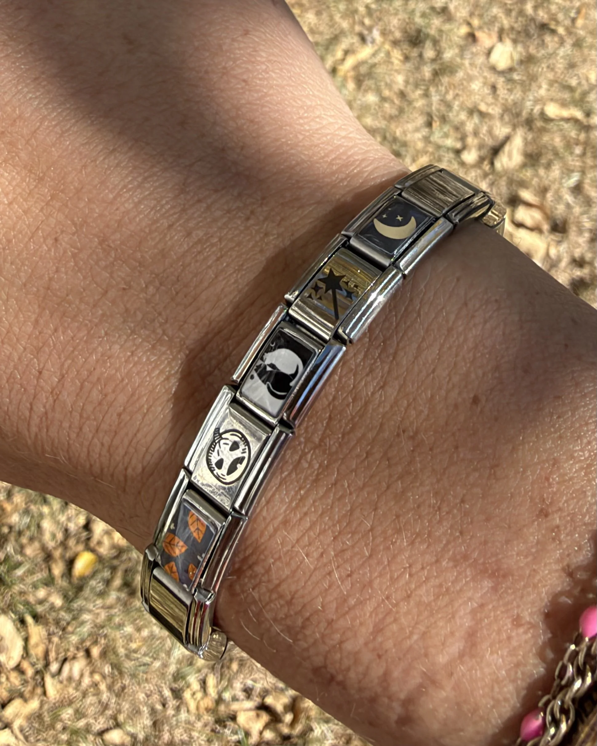 Close-up of a silver Halloween Italian charm bracelet with pumpkin, black cat, wand, and crescent moon designs worn outdoor on a wrist.