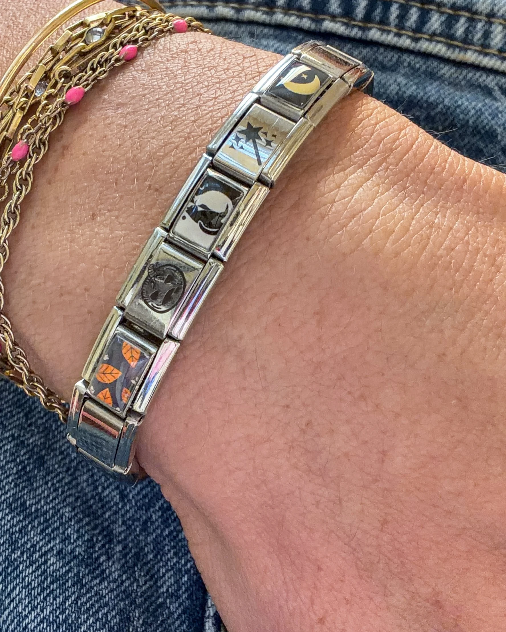 Silver Italian charm bracelet with five Halloween-themed charms worn on a wrist alongside layered gold bracelets.