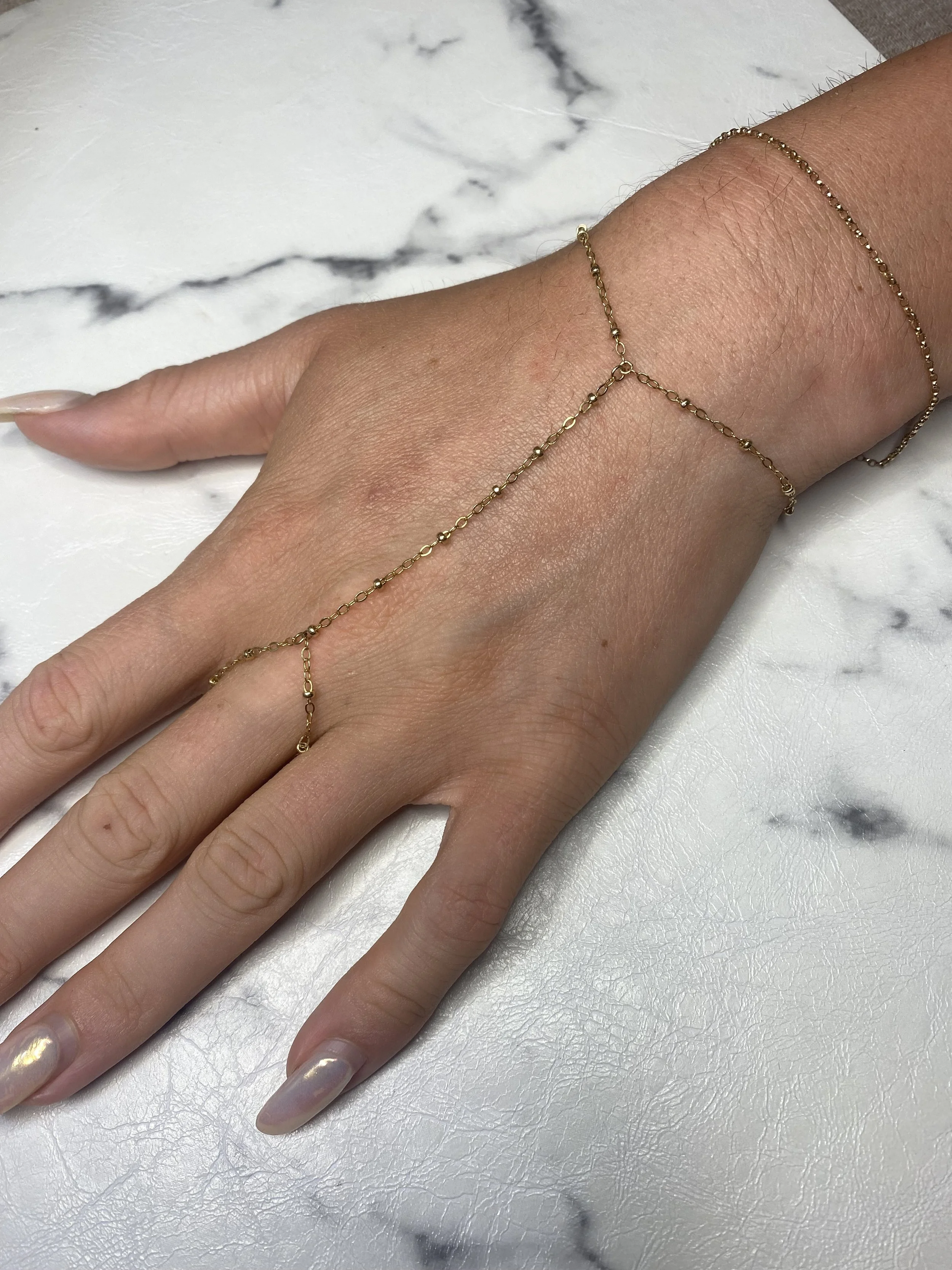 A 14k gold-filled permanent jewelry handchain on a woman's wrist. The chain goes around her wrist like a bracelet, and then has another chain that attaches to a ring around her middle finger.