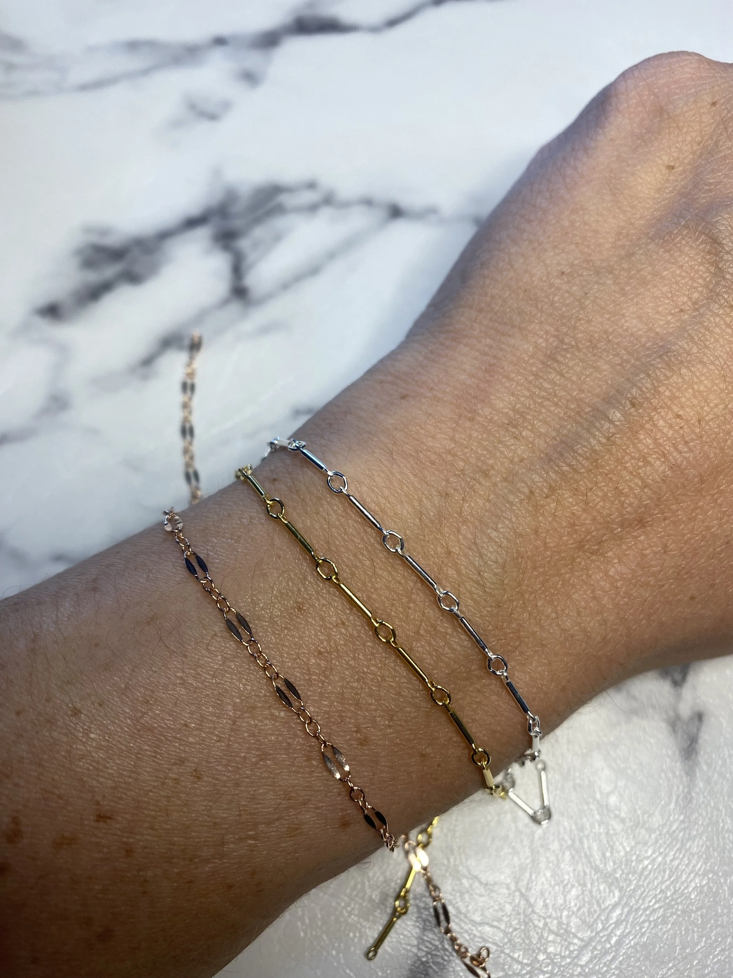 A person's wrist wearing three delicate chain bracelets in gold, silver, and rose gold colors, resting on a white marble surface with gray veining.