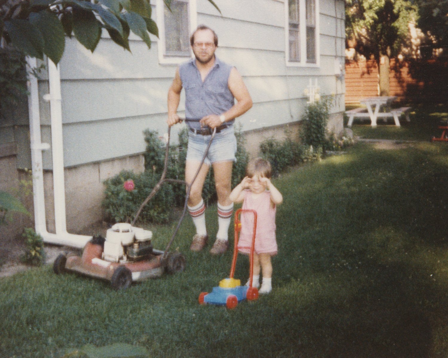 Dad showing Tim how to mow the lawn, 1983 while sporting 'cut-off jeans shorts'.