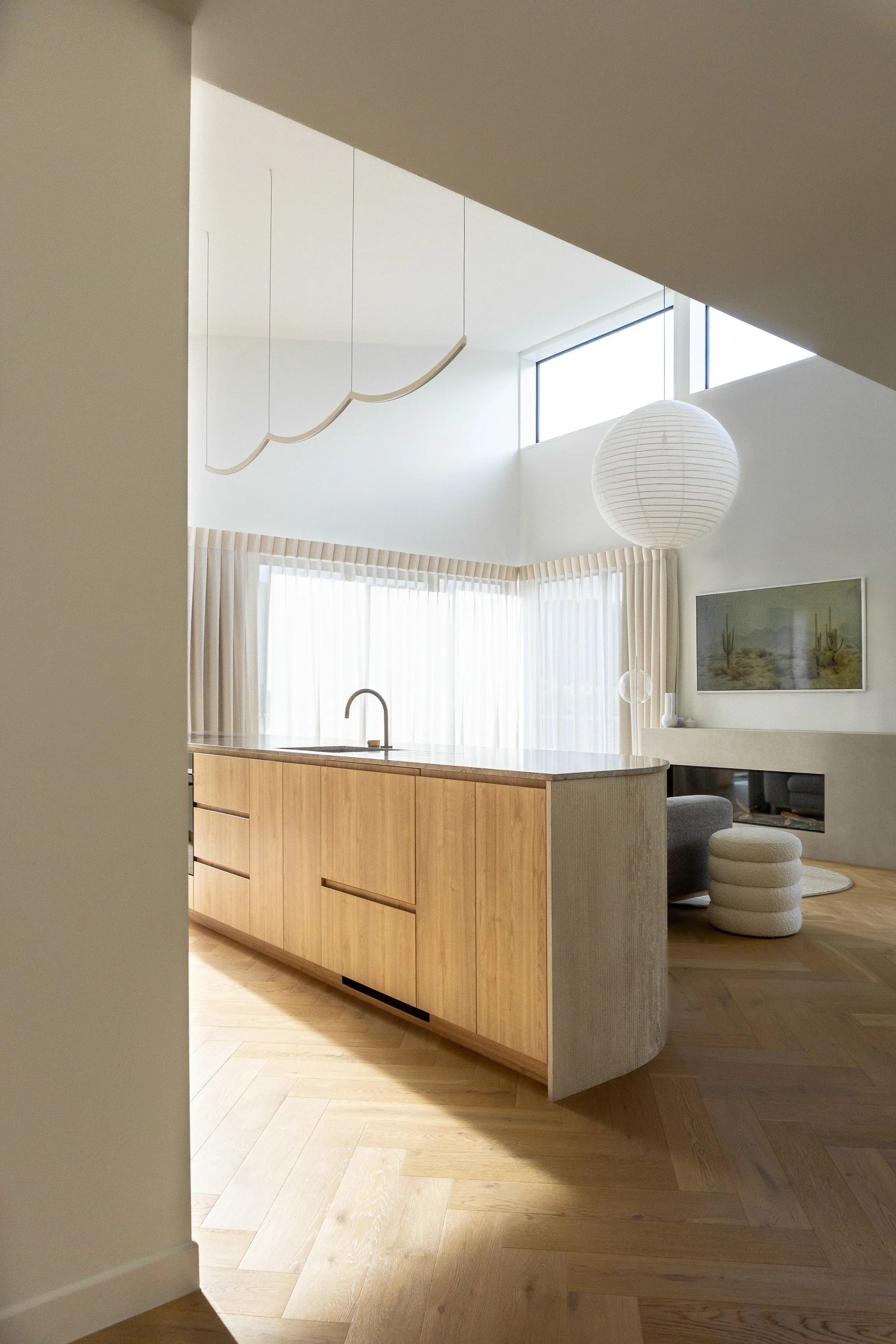 Modern kitchen and living room with minimalist design, wood flooring, white walls, large window with curtains, a large white round paper lantern, a wooden kitchen island with a sink, and a fireplace with framed picture above it.