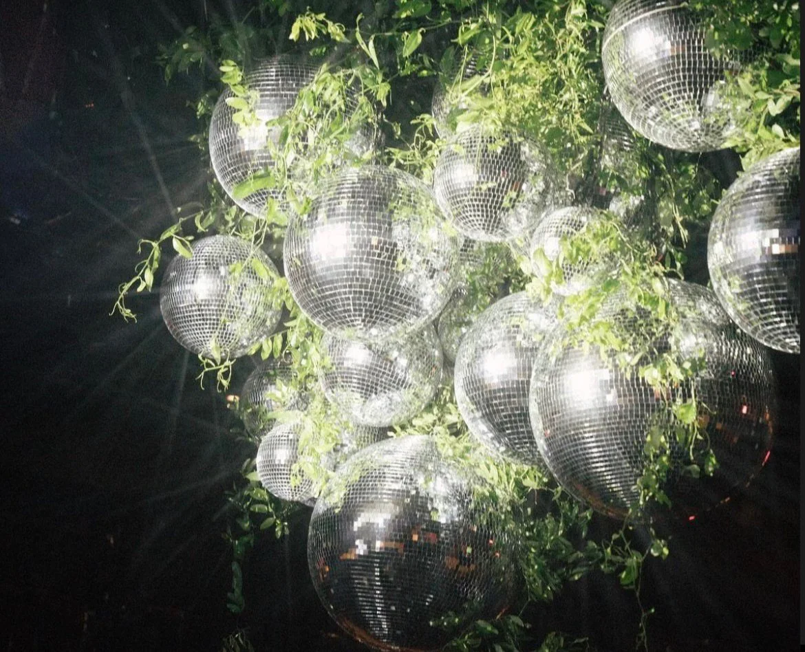 Multiple disco balls hanging among green leaves at night, reflecting light.