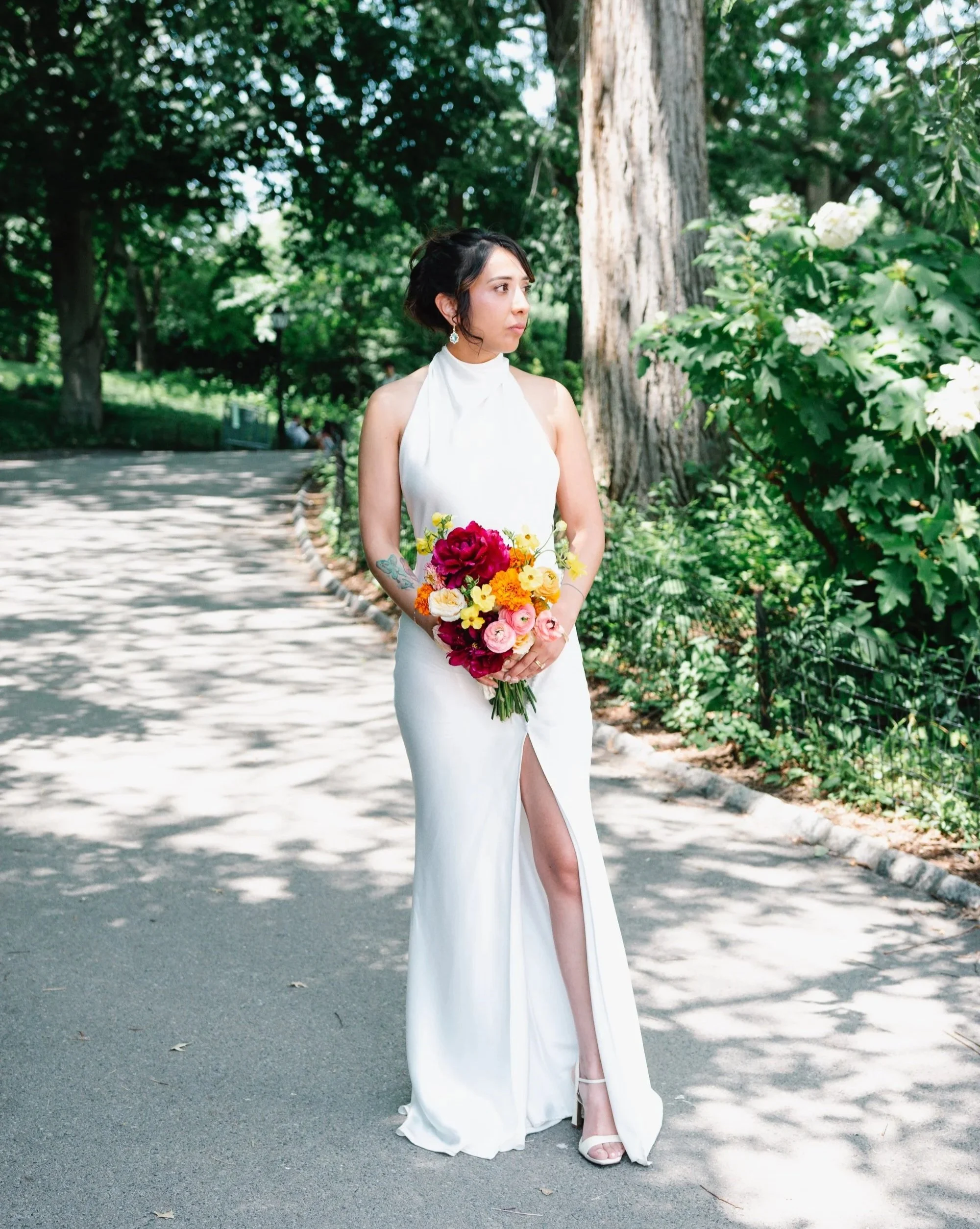 A woman in a white dress holding a bouquet of colorful flowers, standing on a tree-lined path outdoors.