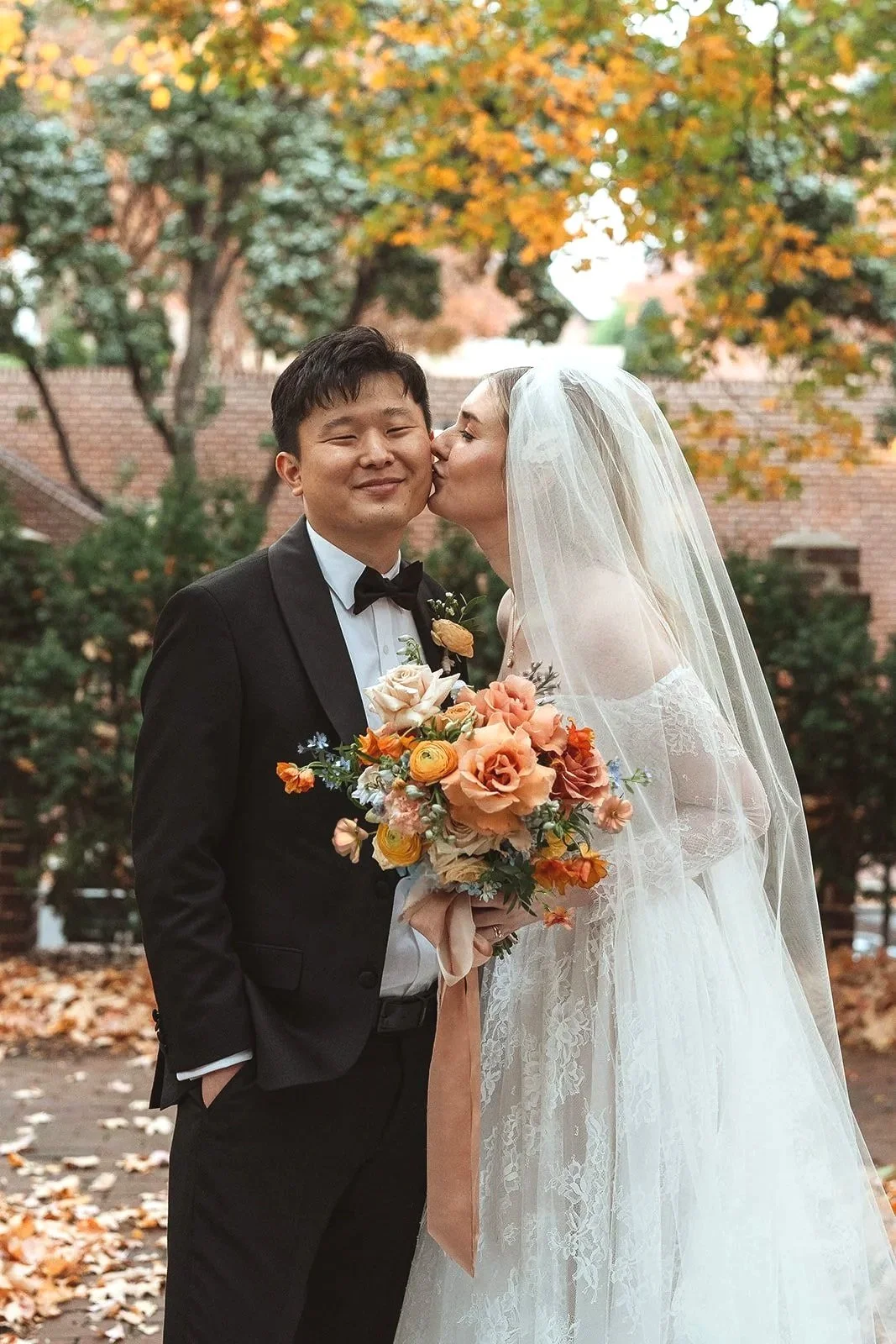 A bride and groom on their wedding day, with the bride kissing the groom on the cheek. The bride is holding a bouquet of orange and peach flowers, and they are standing outdoors with autumn leaves and trees in the background.
