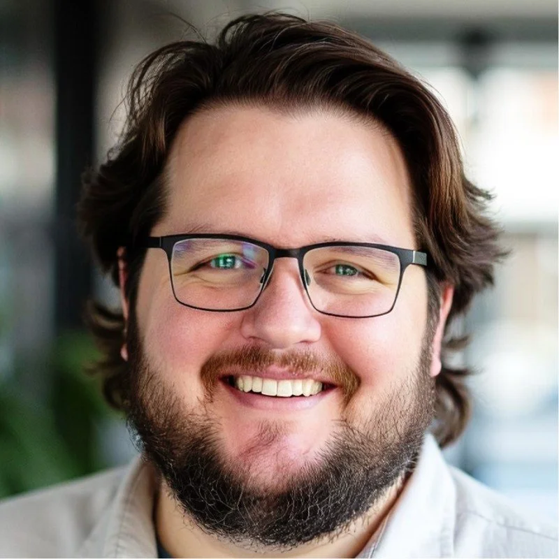 A smiling man with glasses, brown hair, and a beard, in a well-lit indoor setting.