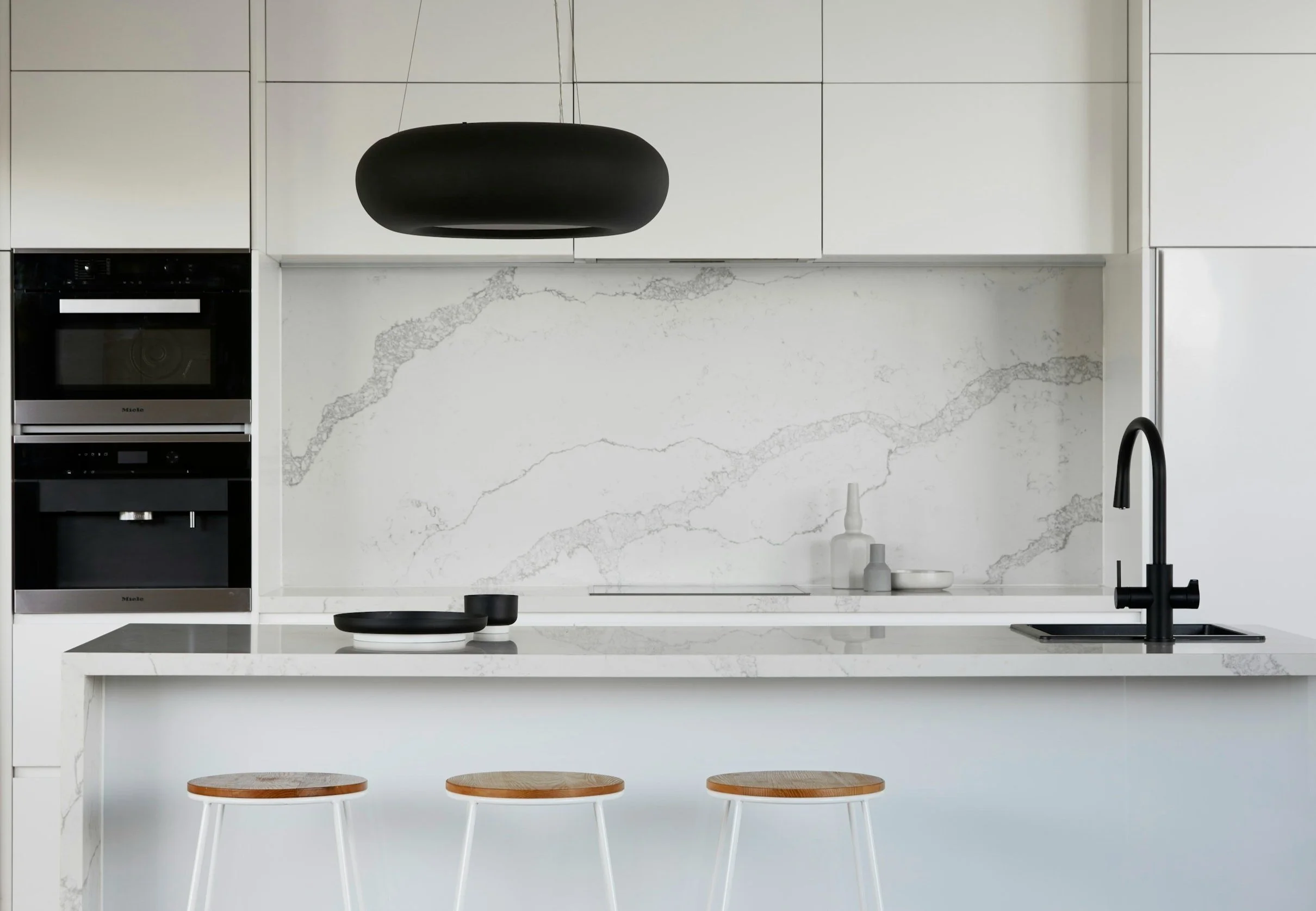 Modern kitchen with white cabinets, black pendant light, marble backsplash, black sink, and wooden bar stools.