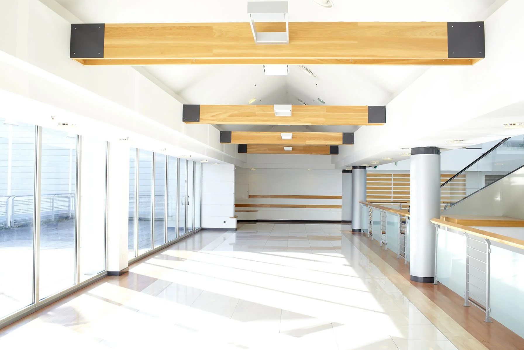 Bright, modern indoor space with large glass windows on the left, white walls, wooden beams on the ceiling, and a polished tile floor.
