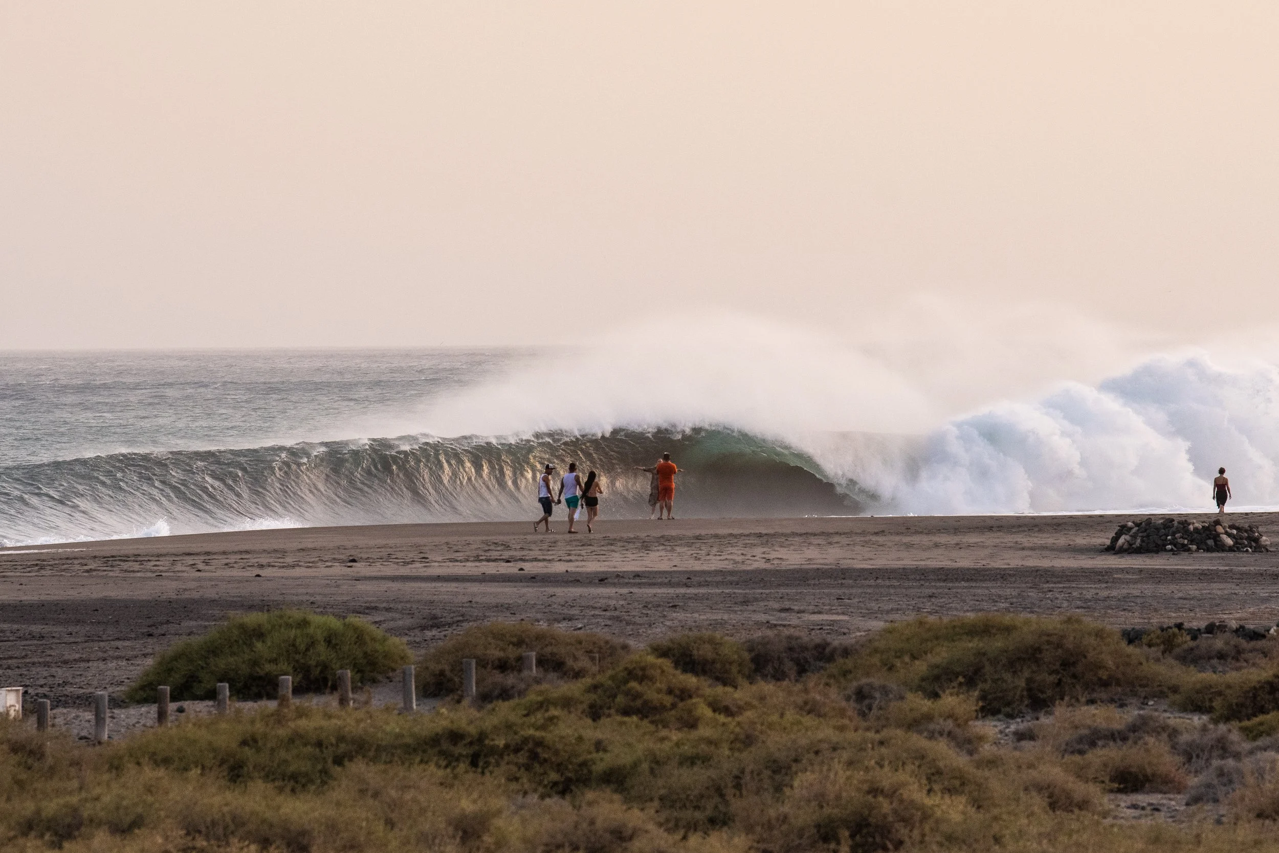 People walking and standing on the beach near large ocean waves during sunset or sunrise.