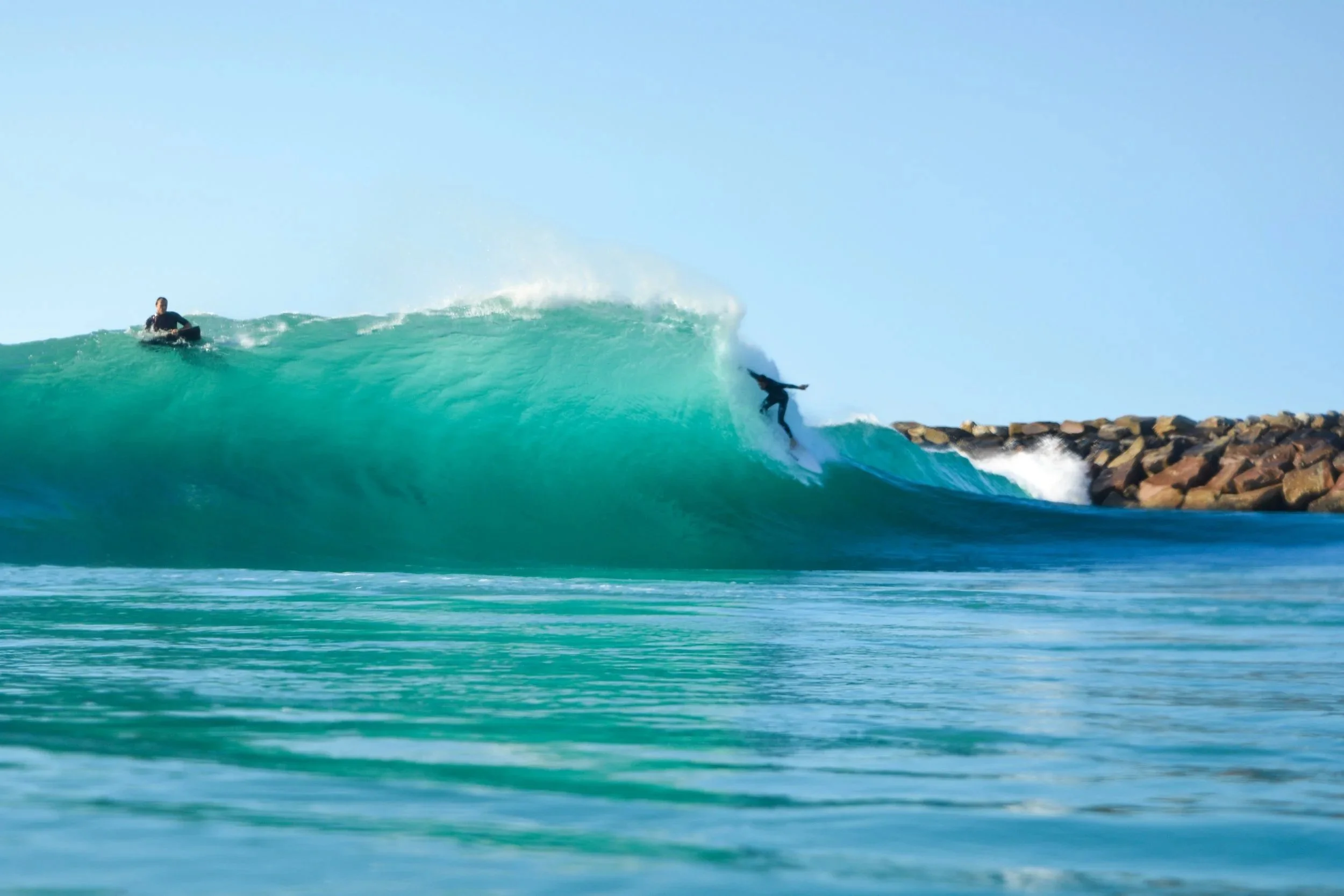 Two surfers riding a large ocean wave near a rocky breakwater, with clear blue sky overhead.