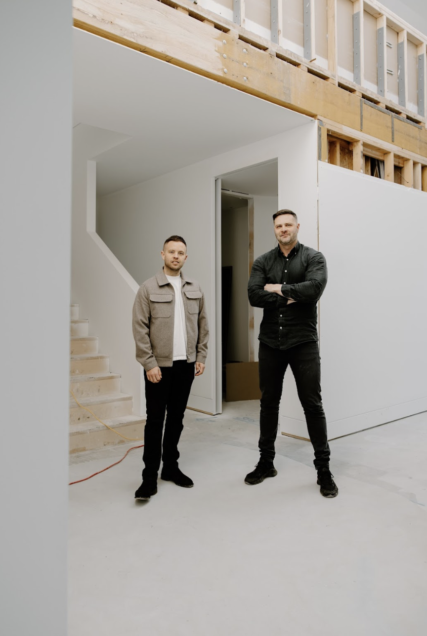 Two men standing inside an unfinished building, one in a beige jacket and the other in a black shirt, with construction materials around.