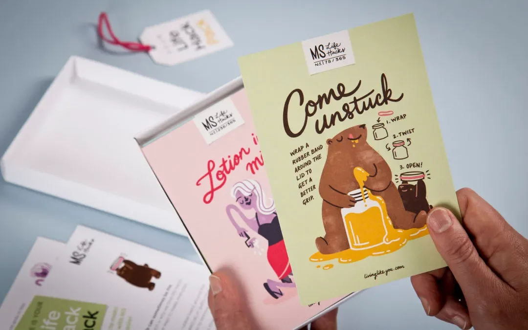 Hand holding an illustrated card with a bear pouring honey from a jar, giving instructions to interfere with packaging, with writing and drawings on a green background. Other similar cards and a white tray are visible.