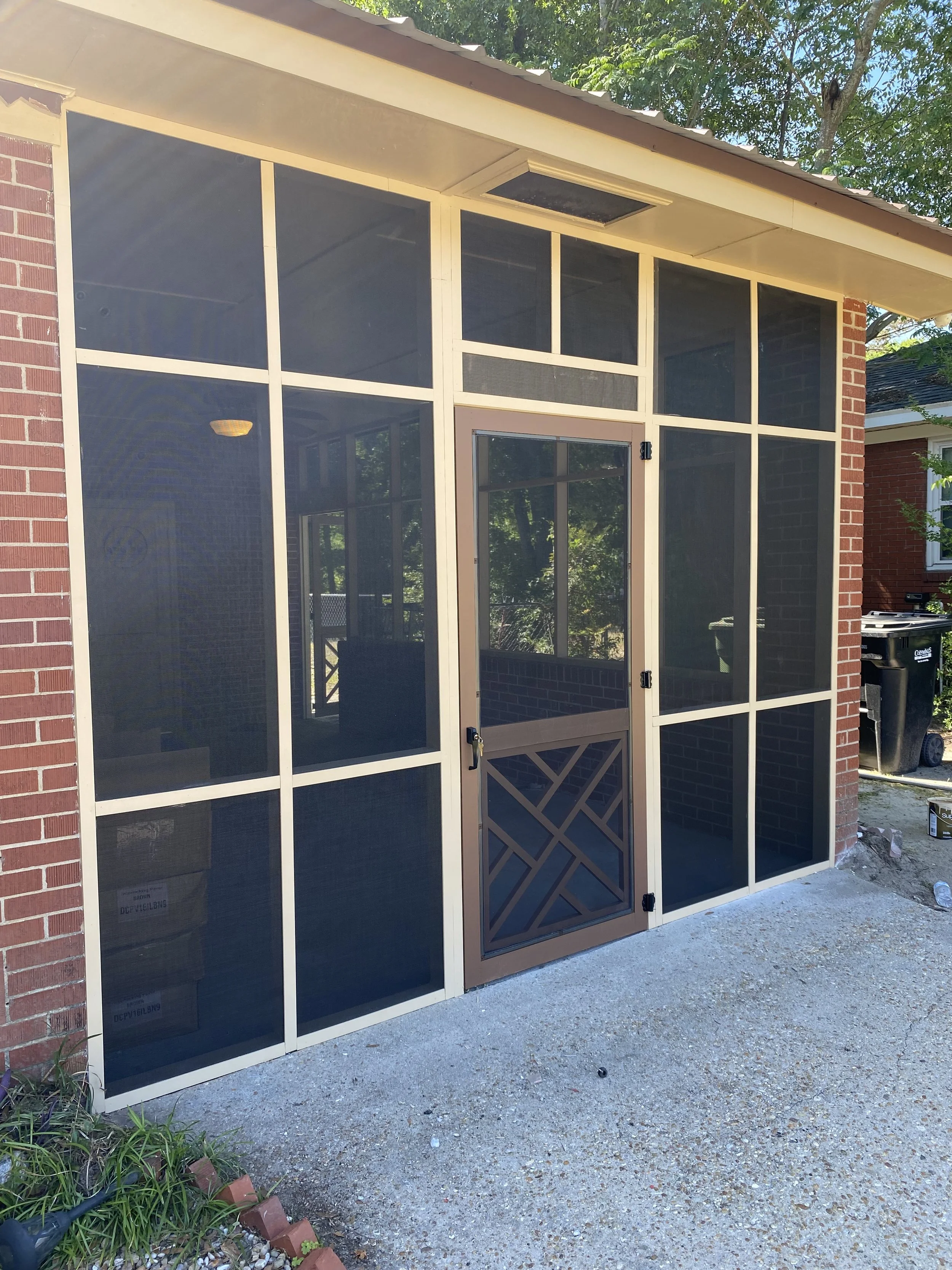 A screened porch with a brick wall on each side, a tan door with decorative pattern, and a metal frame. Green trees are visible above. By Columbus GA Handyman