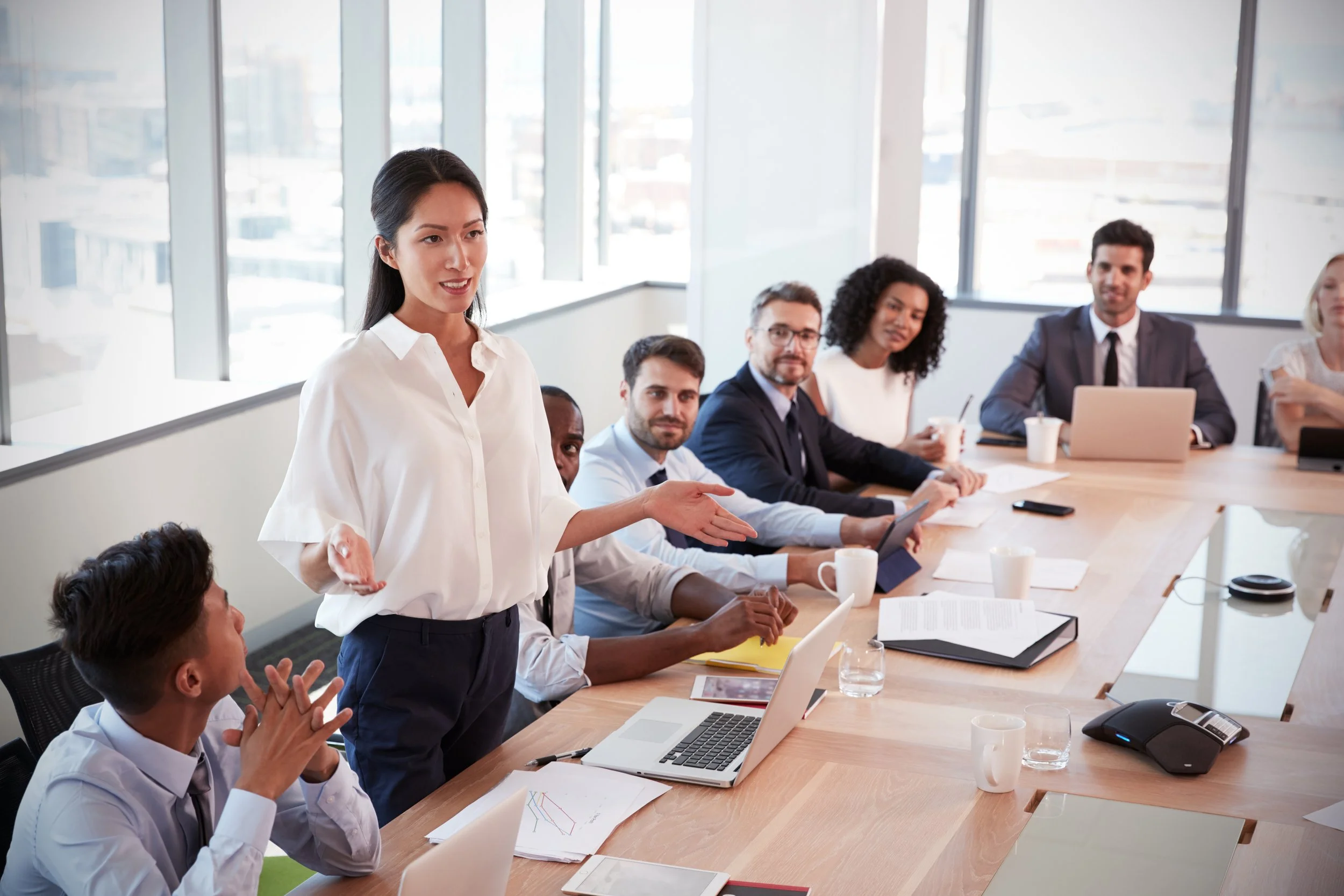 Business meeting with diverse team around a conference table, a woman standing and speaking.