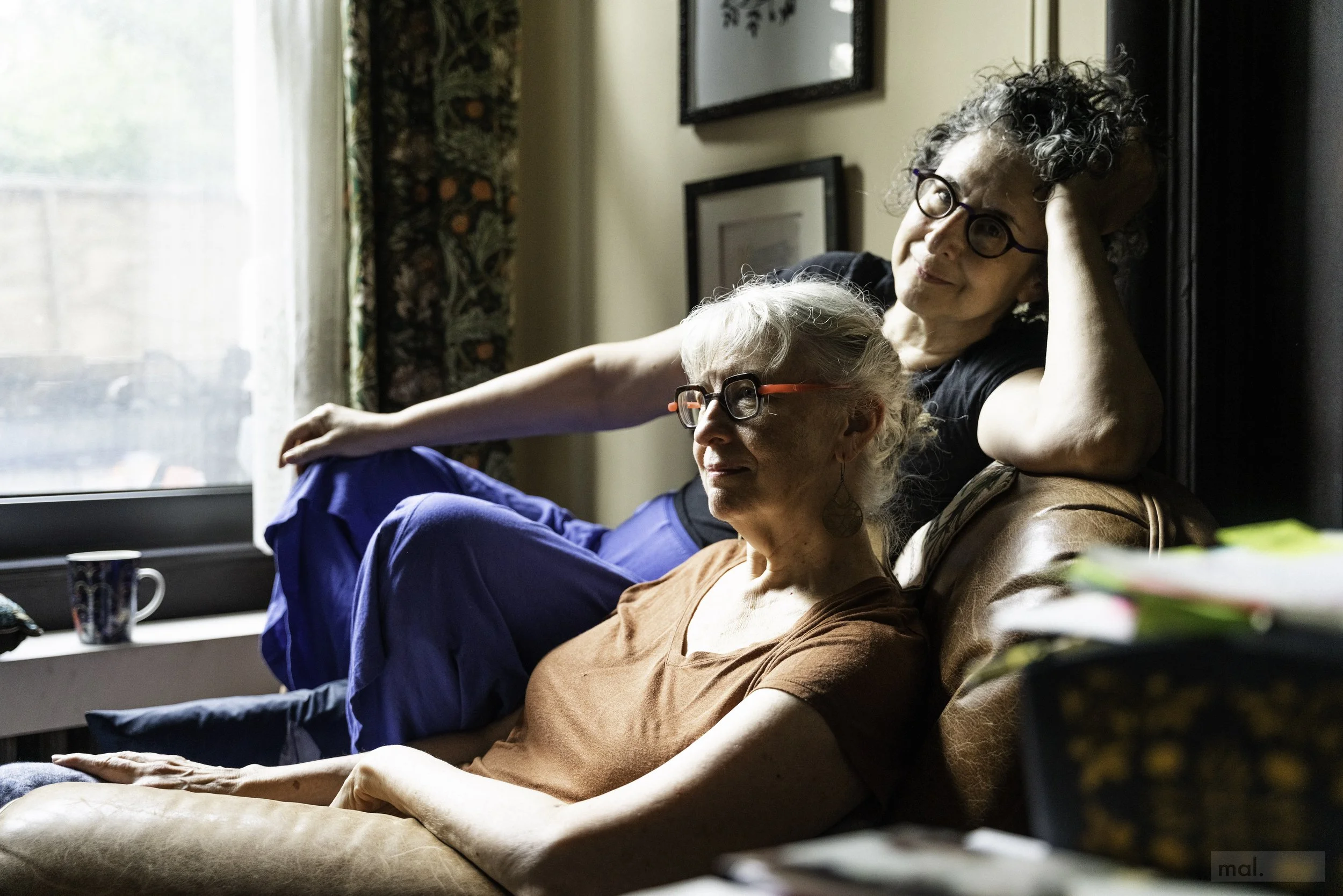 Two women, one elderly and one middle-aged, sitting on a sofa in a living room, both wearing glasses and appearing relaxed and contemplative.