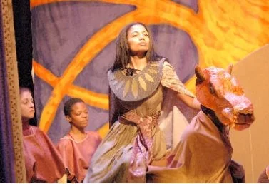 A woman in costume performing on stage with children in the background. One child wears a horse mask.